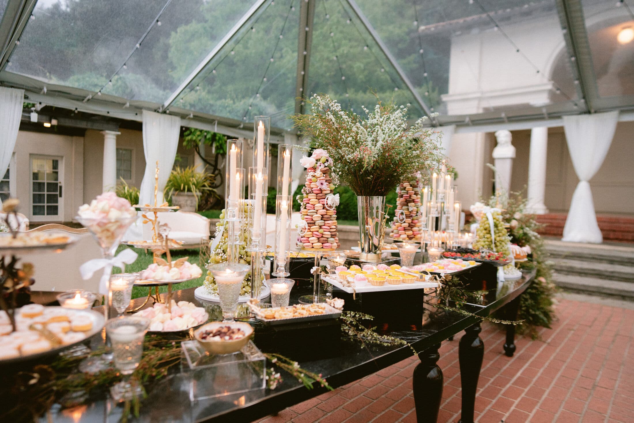 A candlelit black dessert table at dusk under a clear tent, styled with stacked macaron towers, tiered stands, glass dessert cups, and trailing greenery on a brick patio.