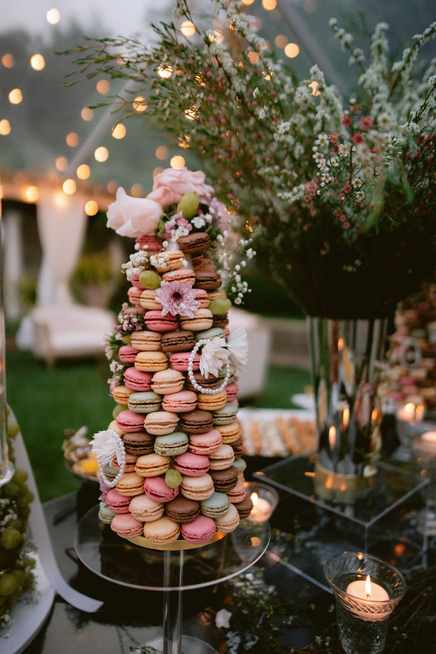 A tall pastel macaron tower dressed with fresh roses, gerbera, and baby's breath, glowing under string lights beside a votive and dried-flower vase on a black garden table.