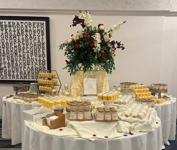 A round white-linen dessert table by Memorable Moments by Ladle n Wok arrayed with rows of layered mousse jars and pudding cups, a labeled menu card, and a tall red-and-white rose centerpiece.