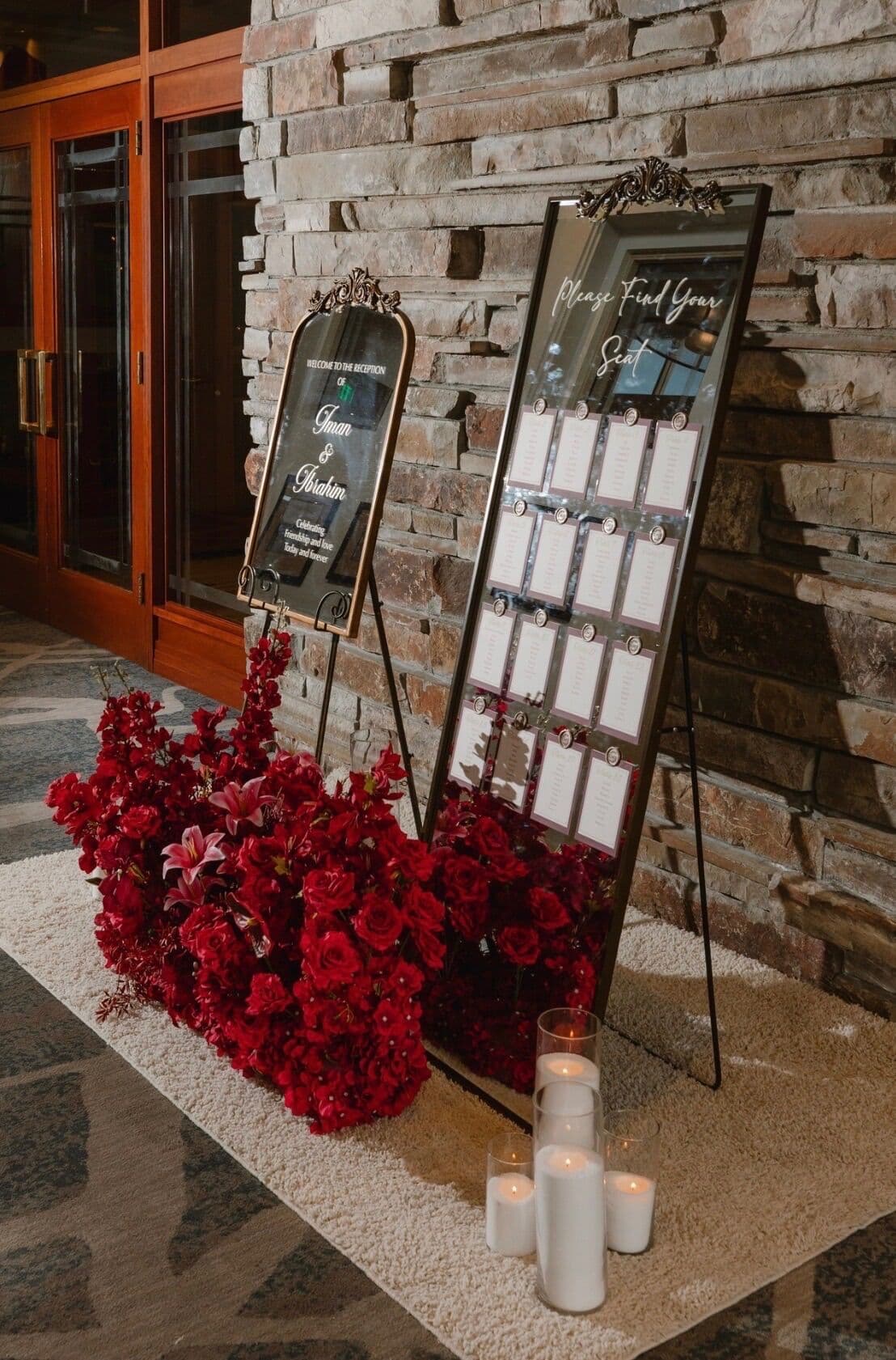 Two black-framed mirror signs on easels against a stacked-stone wall, one a welcome-to-the-reception sign and the other a 'Please Find Your Seat' seating chart with white escort cards, grounded by cascading red roses, lilies and white pillar candles at a Bay Area wedding.