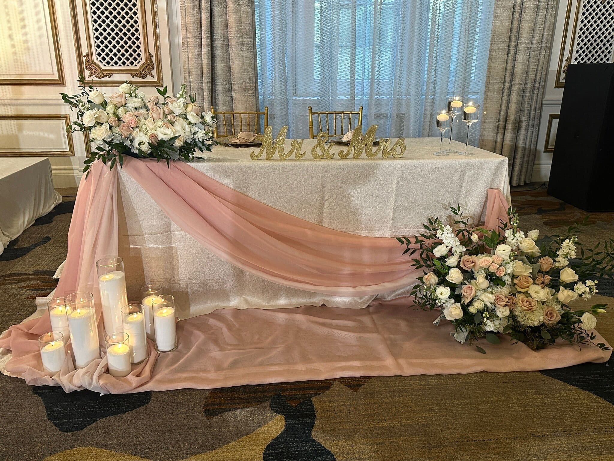 Sweetheart table dressed with a blush organza runner and gold 'Mr & Mrs' letters, framed by a white-and-blush floral arrangement and a row of white pillar candles in glass cylinders at a ballroom wedding.
