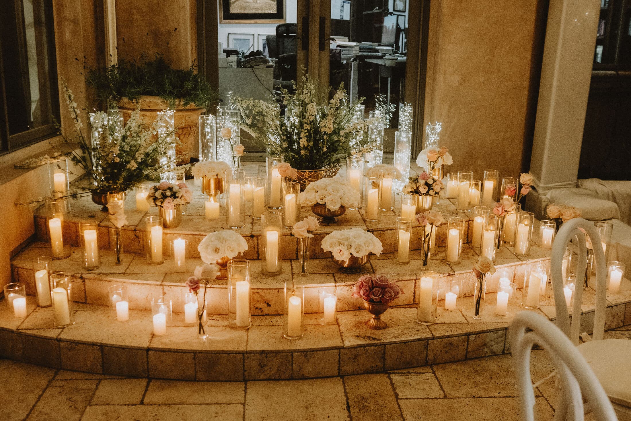 Curved stone steps filled with dozens of white pillar candles interspersed with white and blush floral bouquets, glowing at night beside tall windows at a Bay Area event.