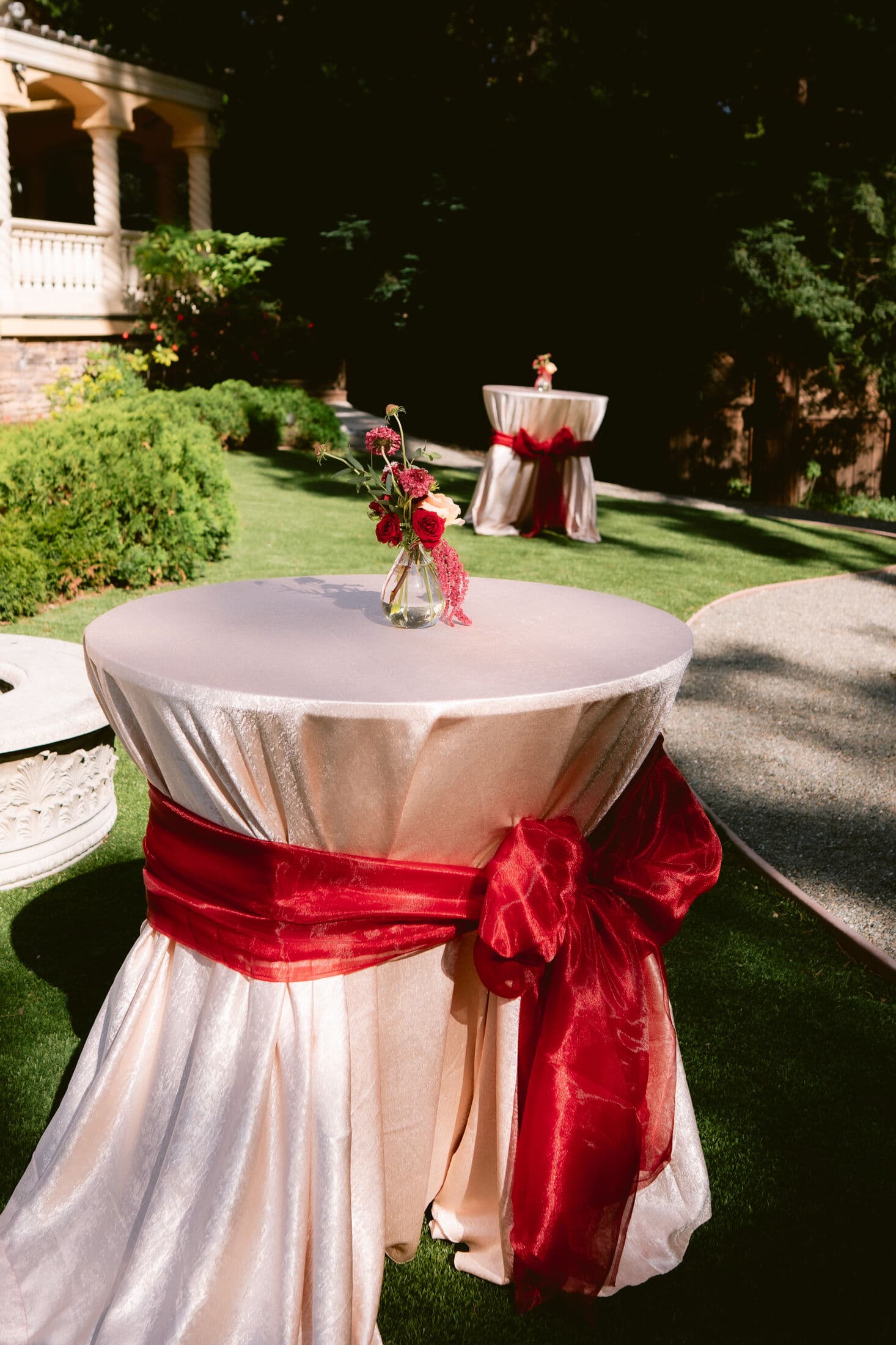 Two round cocktail tables in ivory satin linens cinched with red organza sashes, each holding a small vase of red blooms, set on a manicured garden lawn beside a stone terrace.