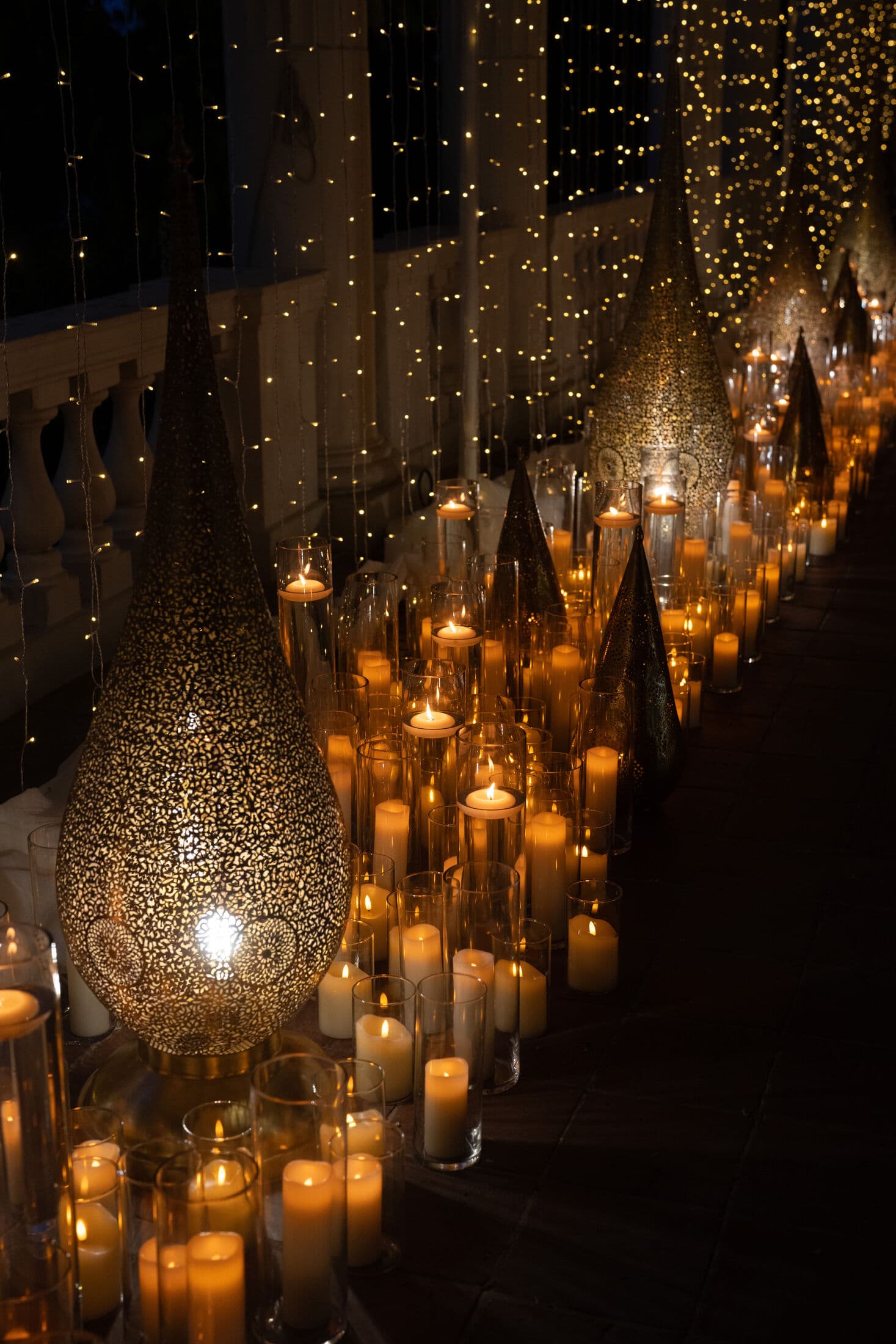 Nighttime terrace lined with pierced-metal gold teardrop lanterns and clusters of white pillar candles against a backdrop of cascading fairy-light curtains at a Bay Area reception.