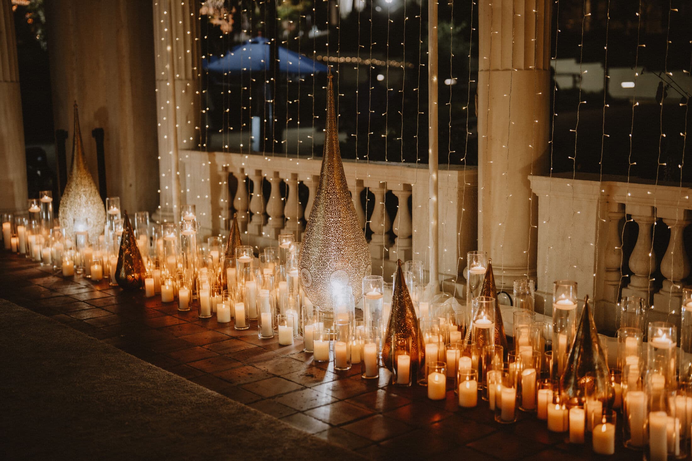 Row of gold filigree cone and teardrop lanterns among massed white pillar candles along a stone balustrade at night, backed by twinkling fairy-light curtains.