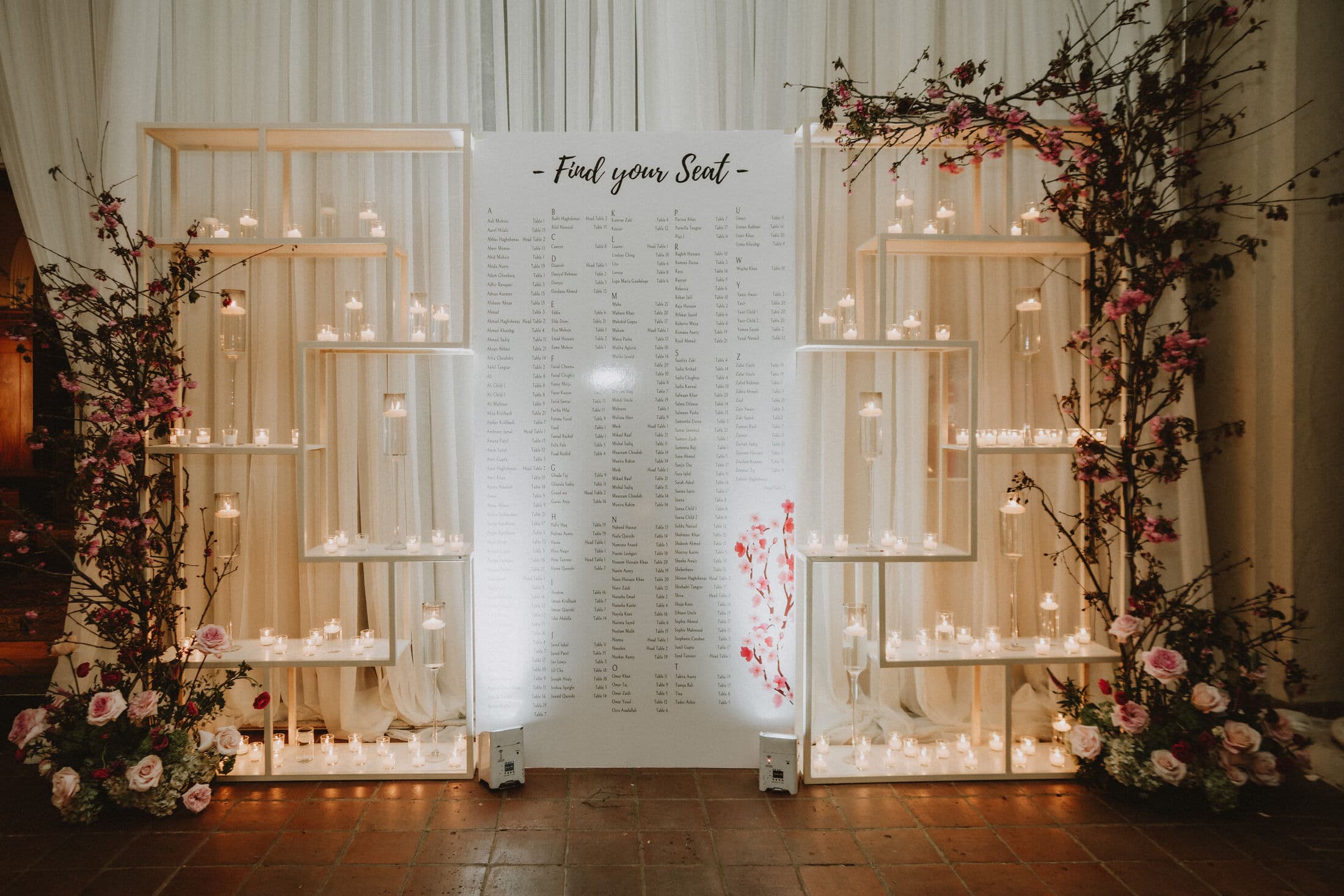 White illuminated 'Find your Seat' seating-chart backdrop flanked by open wooden shelving holding votive candles, framed by climbing pink roses and floral clusters at the base for Memorable Moments by Ladle n Wok.
