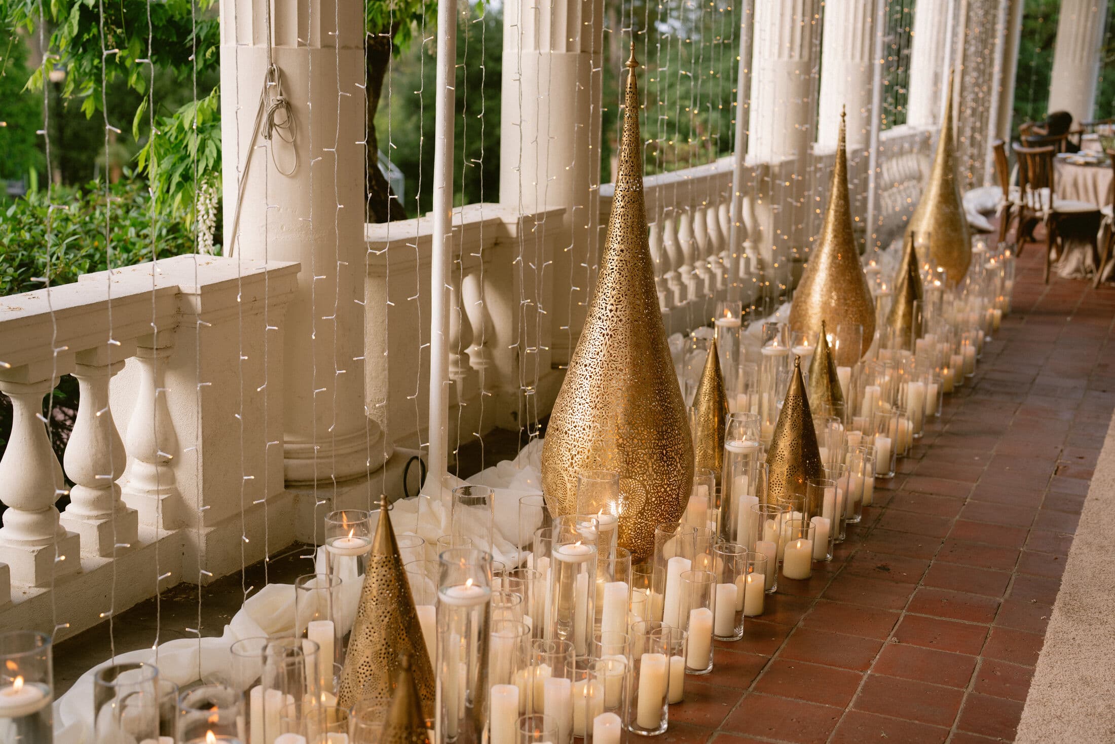 Long verandah edge lined with tall gold perforated teardrop lanterns and white pillar candles in glass cylinders, set against a white stone balustrade and fairy-light curtains at dusk.