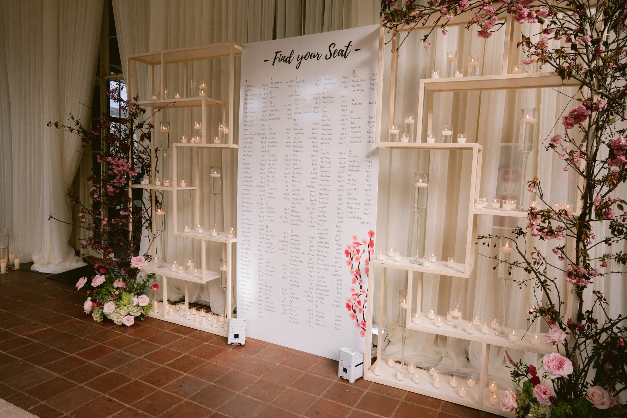 White 'Find your Seat' seating-chart panel set between two wooden geometric shelving units filled with votive candles and pale escort cards, dressed with cherry blossom branches and blush roses at a Bay Area wedding.