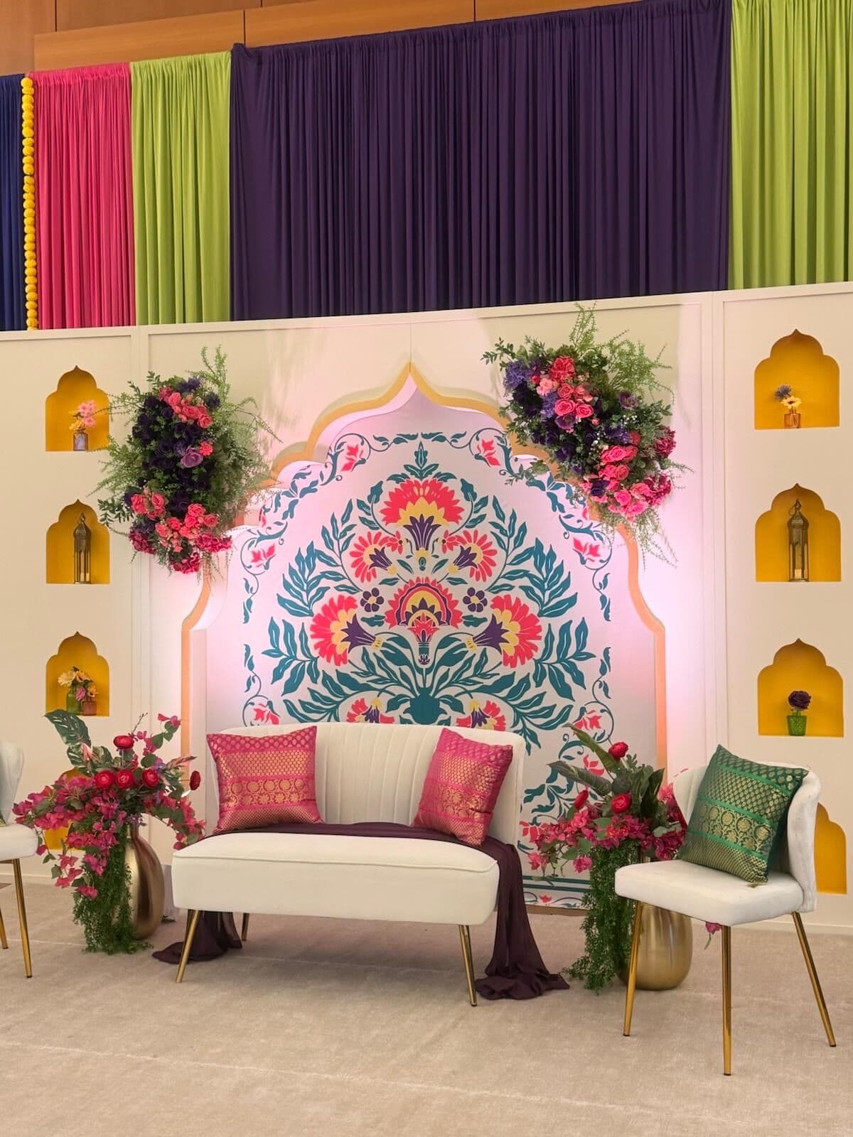 Mehndi stage with an arched backdrop hand-painted in teal, coral and magenta floral motifs, flanked by ivory walls with gold arched niches holding lanterns; a white velvet loveseat with pink brocade cushions sits beneath jewel-toned magenta, green and purple drapery.