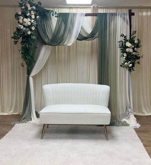Wedding backdrop of sage green and ivory sheer drapery swagged over a bar, with white rose and eucalyptus clusters at the corners and a channel-tufted white loveseat on a plush rug.