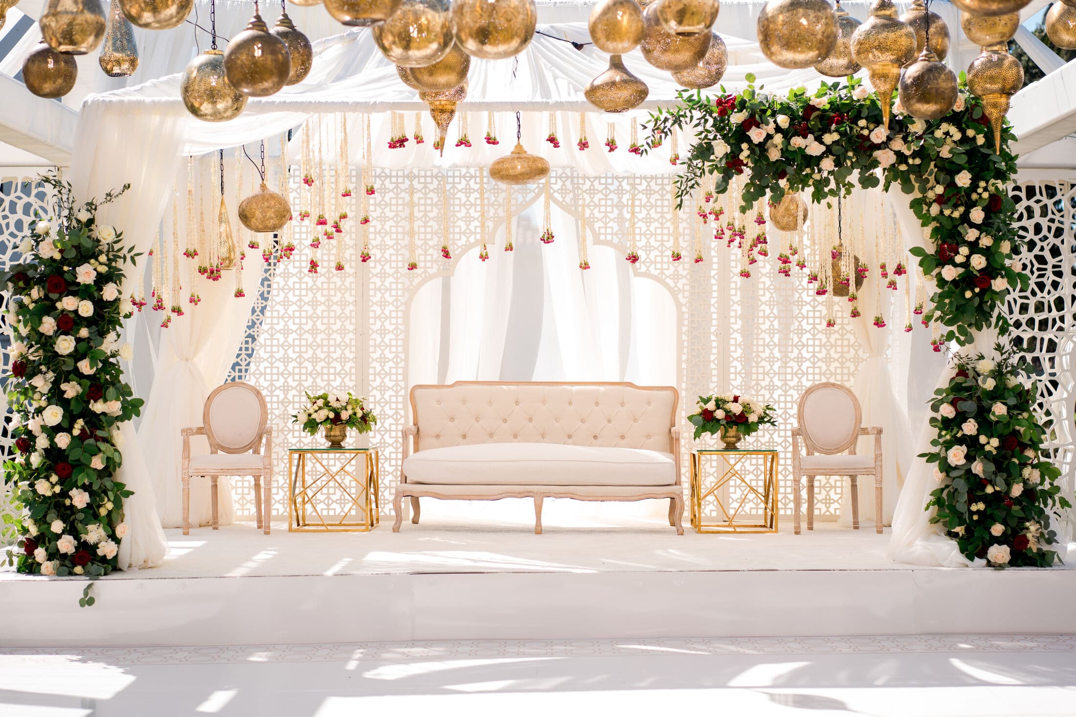 Outdoor mandap-style walima stage in ivory with white lattice screens, greenery-and-rose garlands, gold hanging orbs and strings of blush florals over a tufted cream settee flanked by upholstered oval-back chairs and gold side tables.