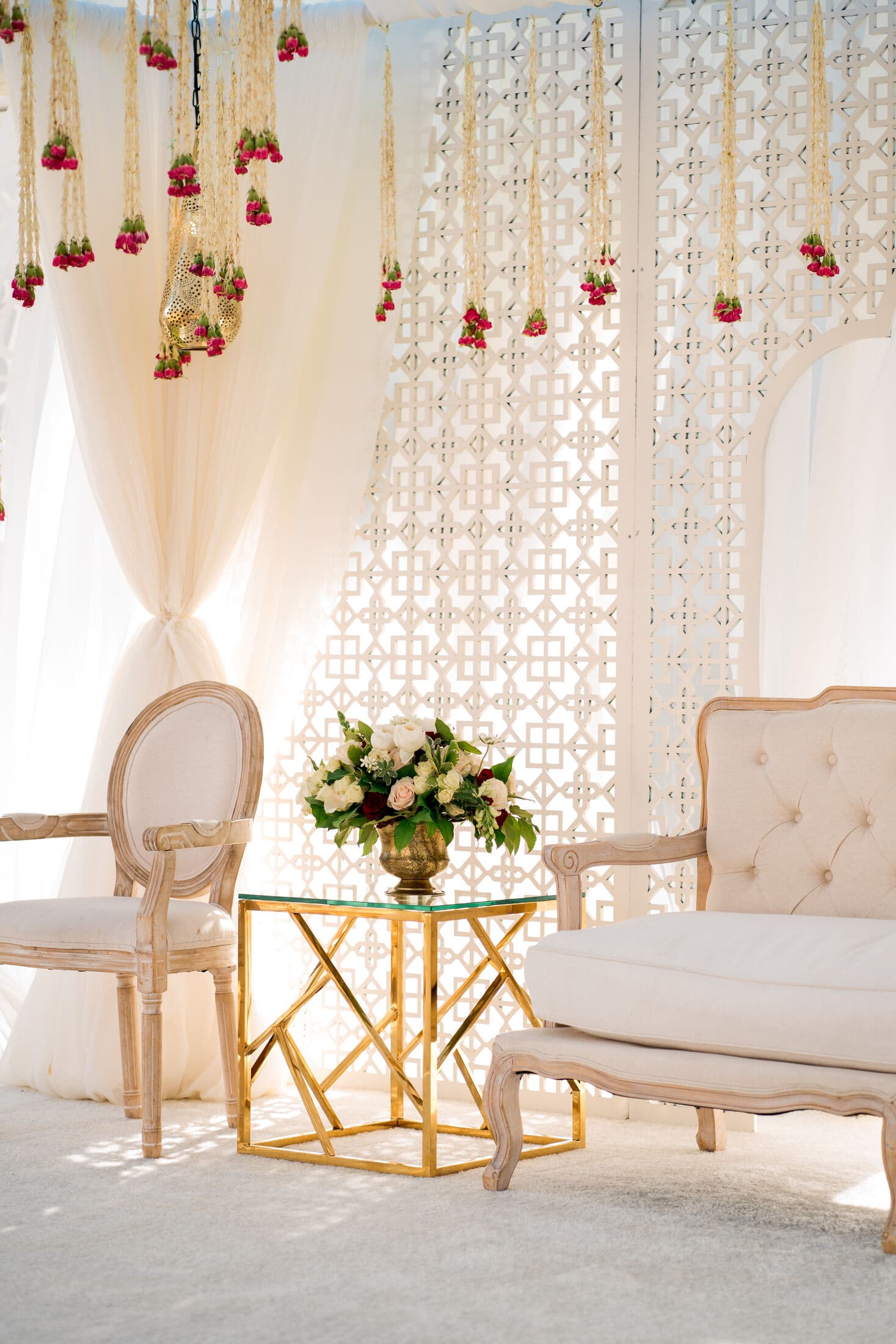 Detail of an ivory wedding stage with a white geometric lattice screen, sheer draping and hanging strands of marigold and red rose garlands, set with a tufted cream settee, a light oval-back chair and a gold-framed glass side table holding white roses.
