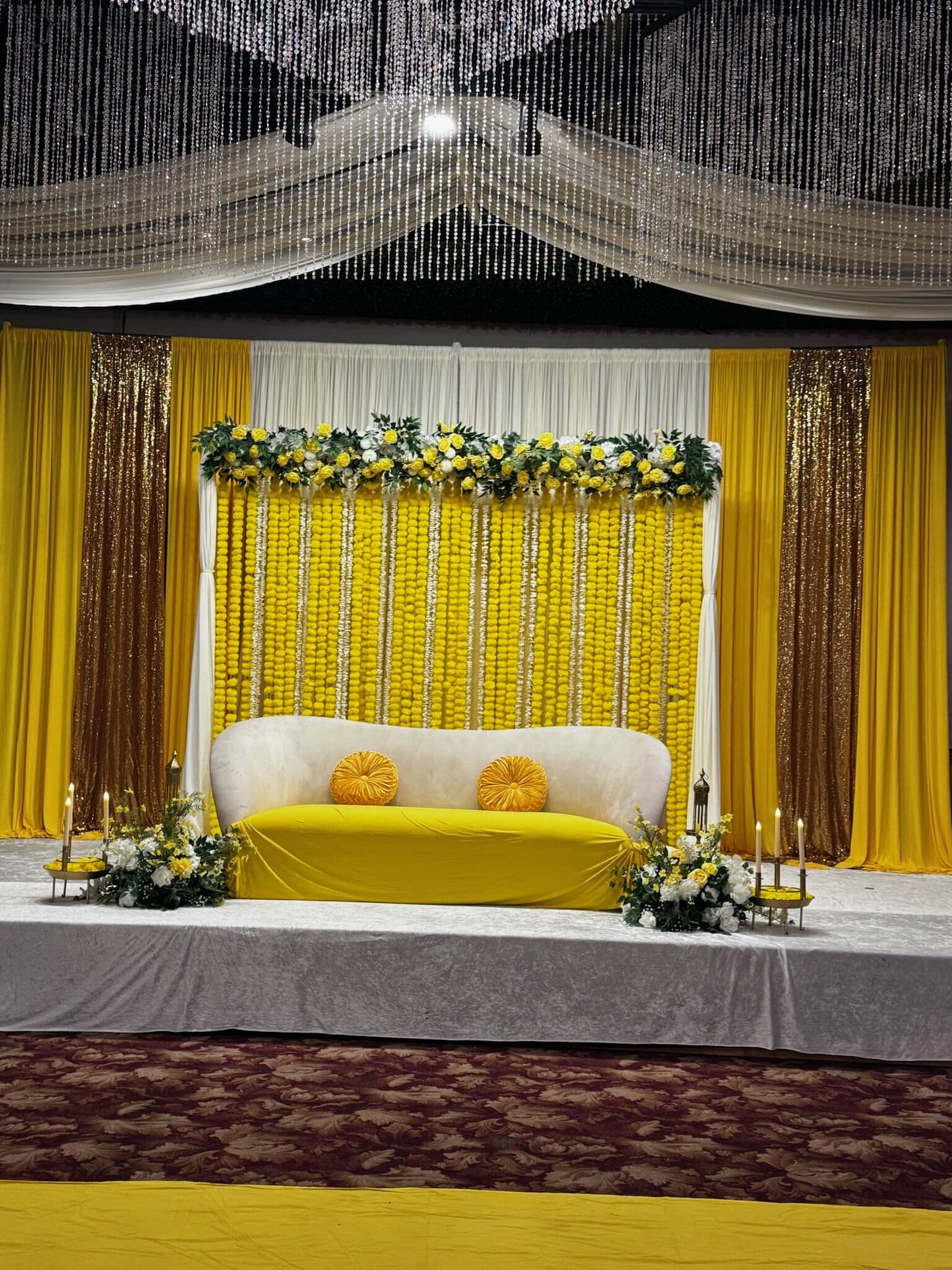 Mehndi stage with a bright yellow backdrop of hanging marigold-style garlands framed by gold sequin and yellow drapery under a swagged crystal-beaded canopy; a cream tufted sofa with yellow accents sits on a yellow-draped platform.