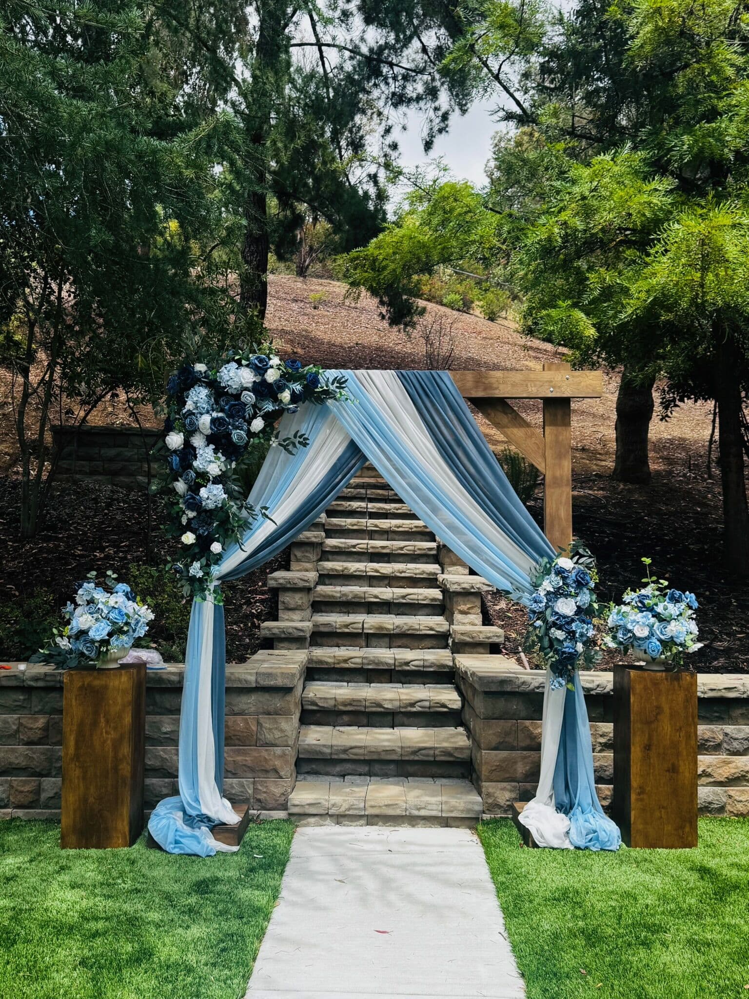 Outdoor Bay Area hillside ceremony arch draped in dusty blue and white chiffon with blue and white rose clusters, set on wood pedestals over a stone staircase and vineyard, along a white aisle runner.