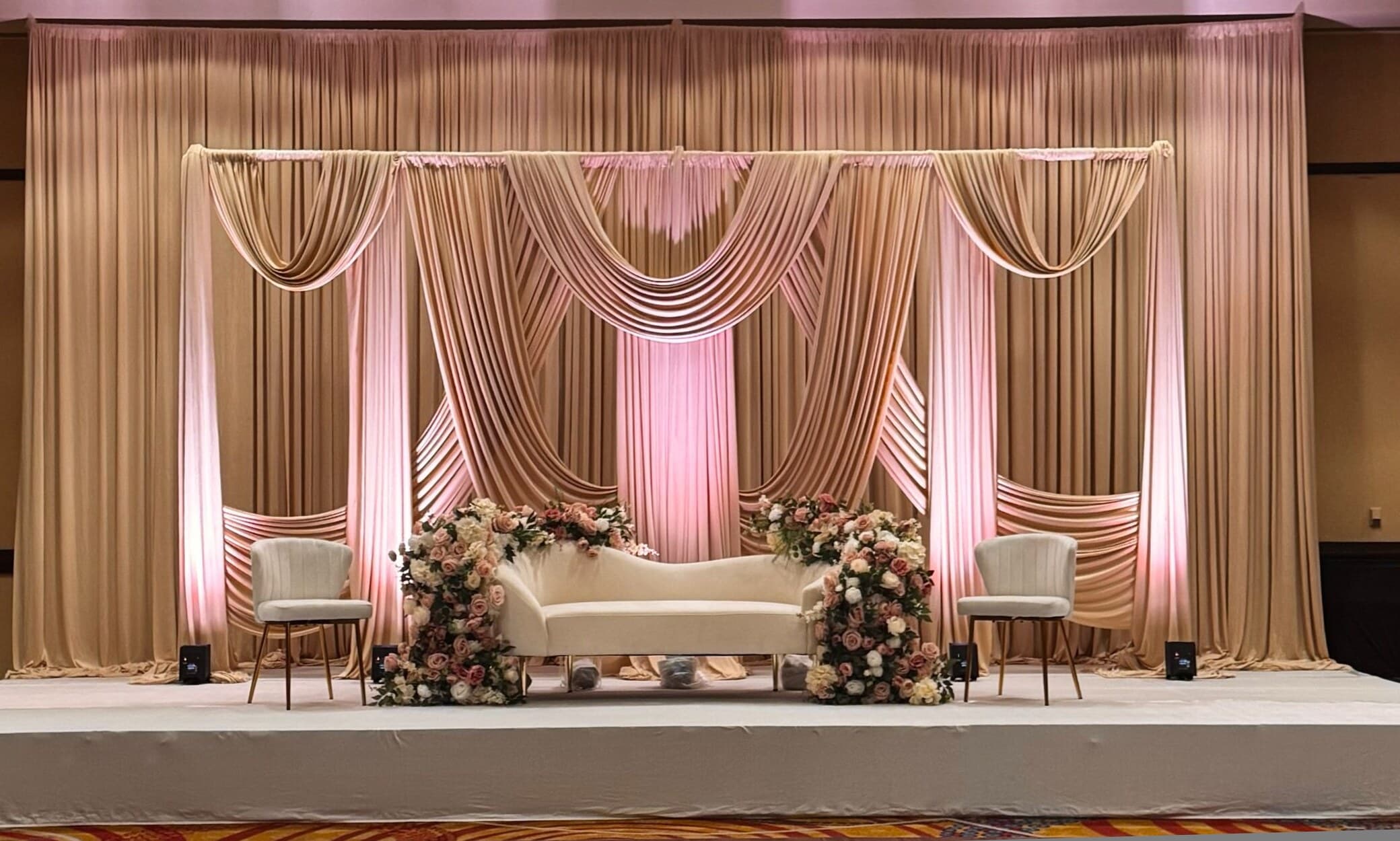 Banquet-hall wedding stage with layered champagne and blush pleated drapery lit in soft pink, a curved ivory sofa centered between mixed blush floral arrangements and two accent chairs on a raised platform.