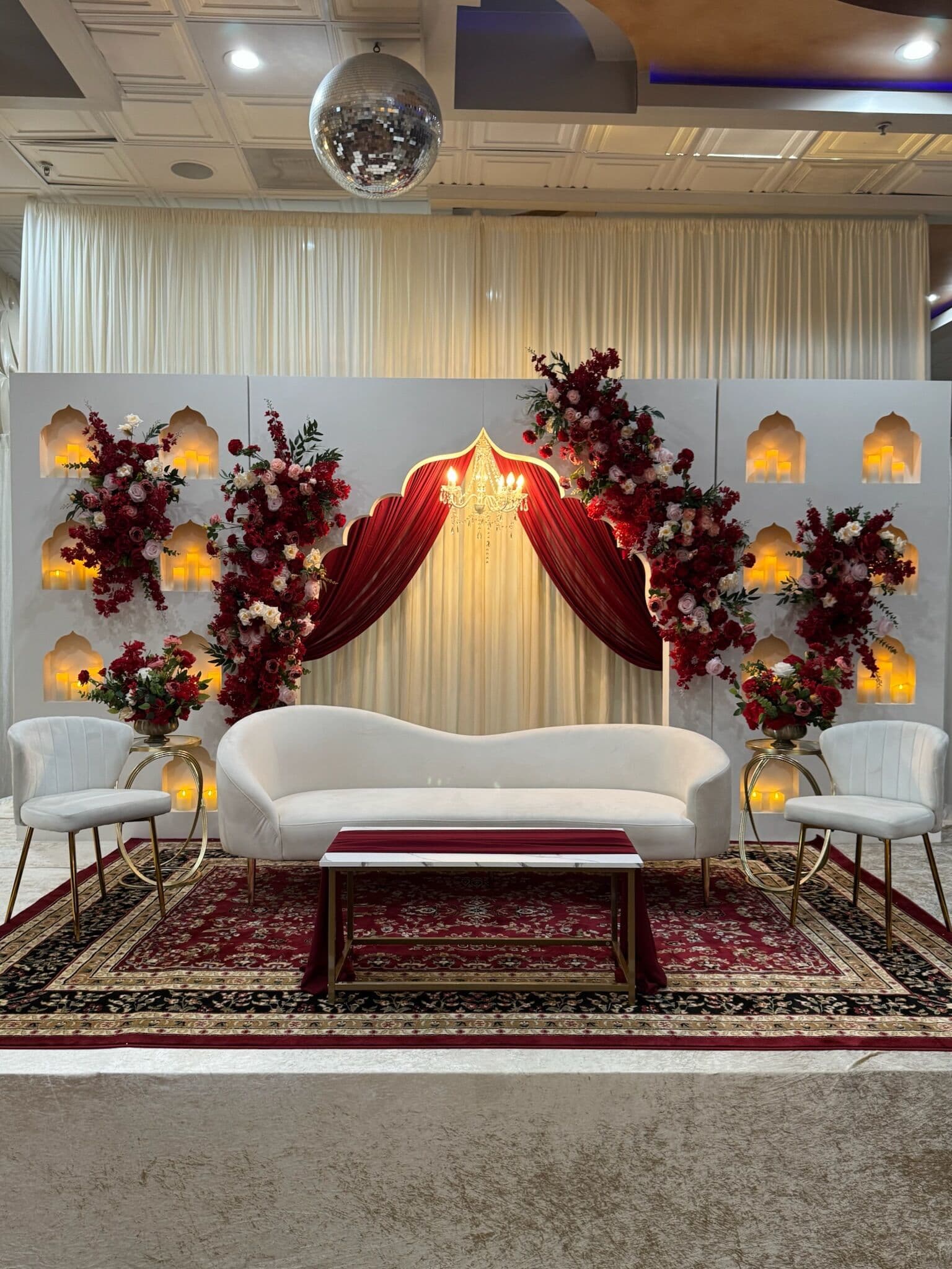 Walima stage with a white backdrop, gold arched wall niches lit warm, deep burgundy velvet swag and lush burgundy-and-white rose arrangements framing a curved white sofa on an ornate red patterned rug, with a disco ball overhead.