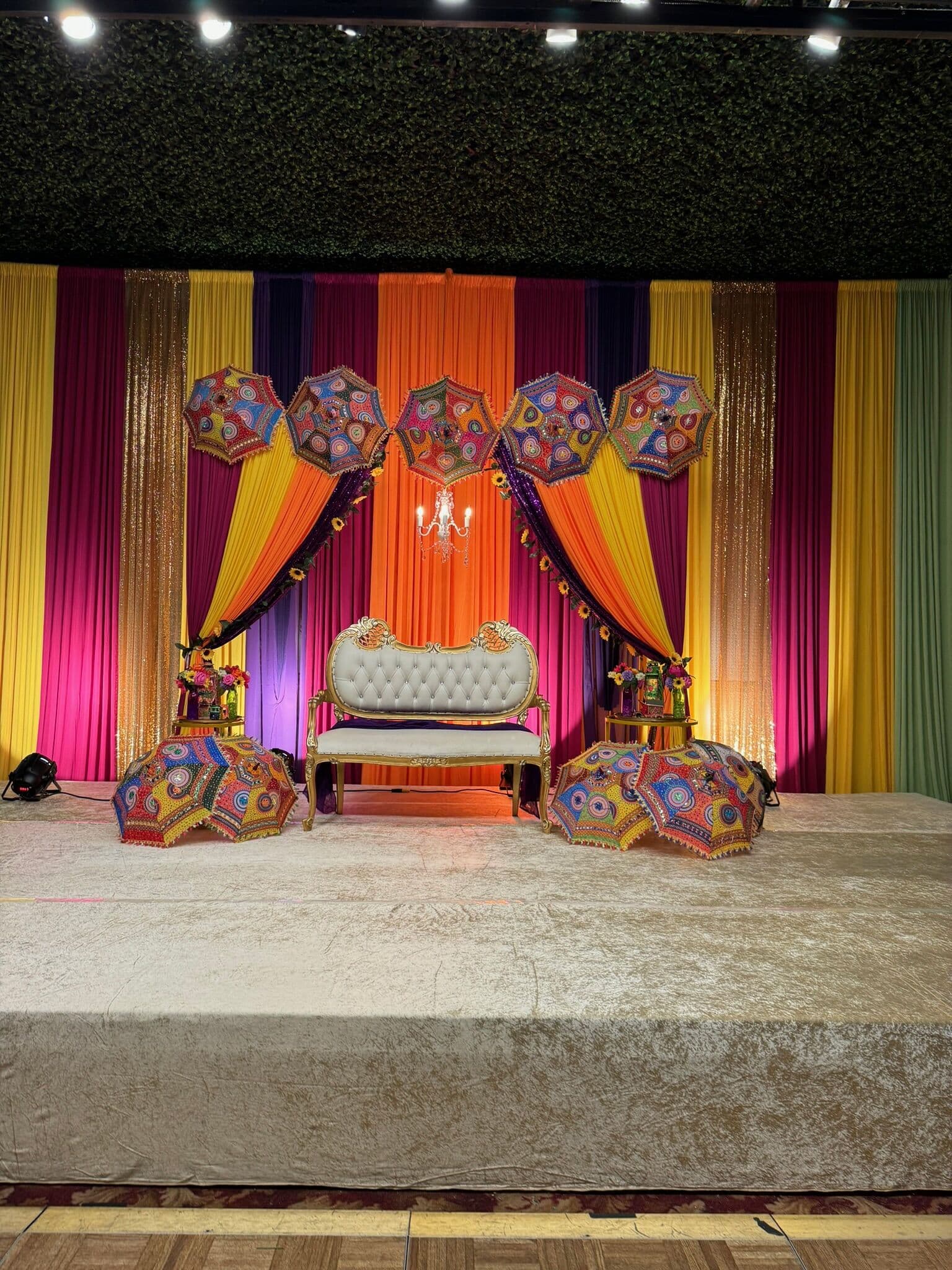 Colorful mehndi stage with rainbow-striped drapery in pink, yellow, orange and gold, decorated with embroidered Rajasthani umbrellas, a small chandelier and a tufted ivory settee on a raised platform.