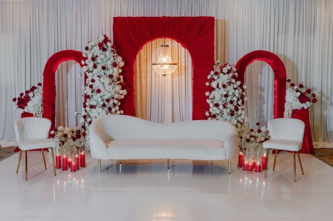 Symmetrical wedding stage with three red ruched arches against ivory drapery, a crystal chandelier, cascading white and burgundy rose columns, a curved white sofa and accent chairs framed by red pillar candles.