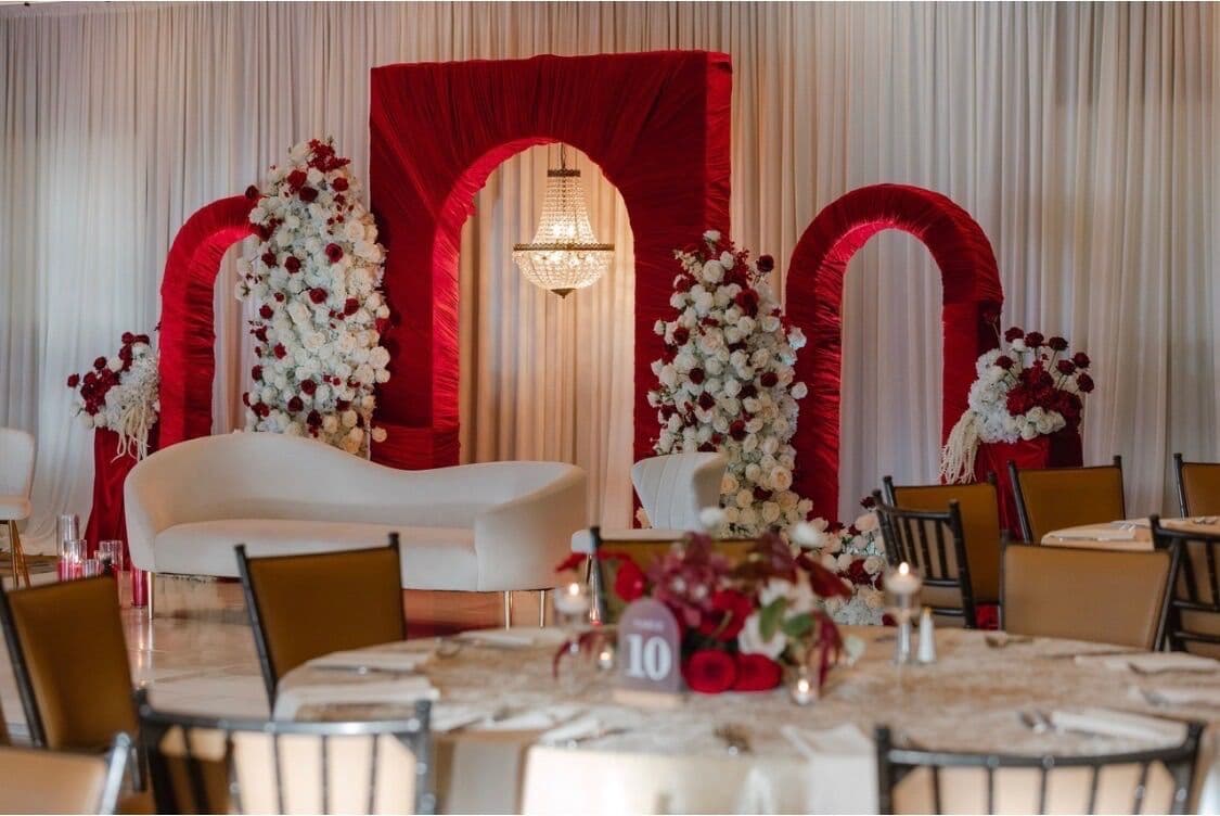 Reception ballroom by Memorable Moments by Ladle n Wok with a red ruched triple-arch stage, crystal chandelier and white-and-burgundy rose columns behind a curved ivory sofa, viewed past round guest tables with red centerpieces.