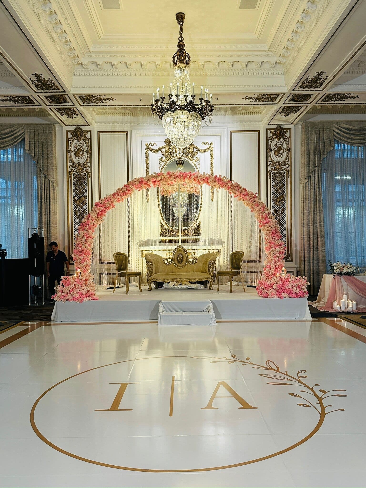 Grand ballroom walima stage with a full pink-and-blush rose arch over a gold tufted settee and gold chairs on a white platform, set in an ornate room with a crystal chandelier and a monogrammed white dance floor.