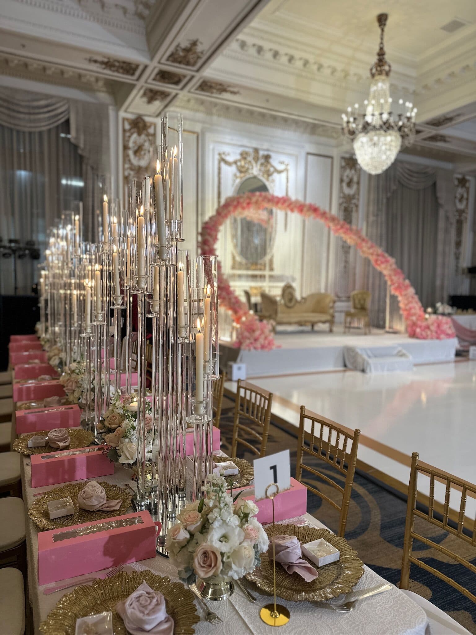Ballroom reception view of a blush rose arch and gold seating on a white dance floor, seen along a head table lined with tall crystal candelabra, gold charger plates and pink risers with low blush centerpieces.