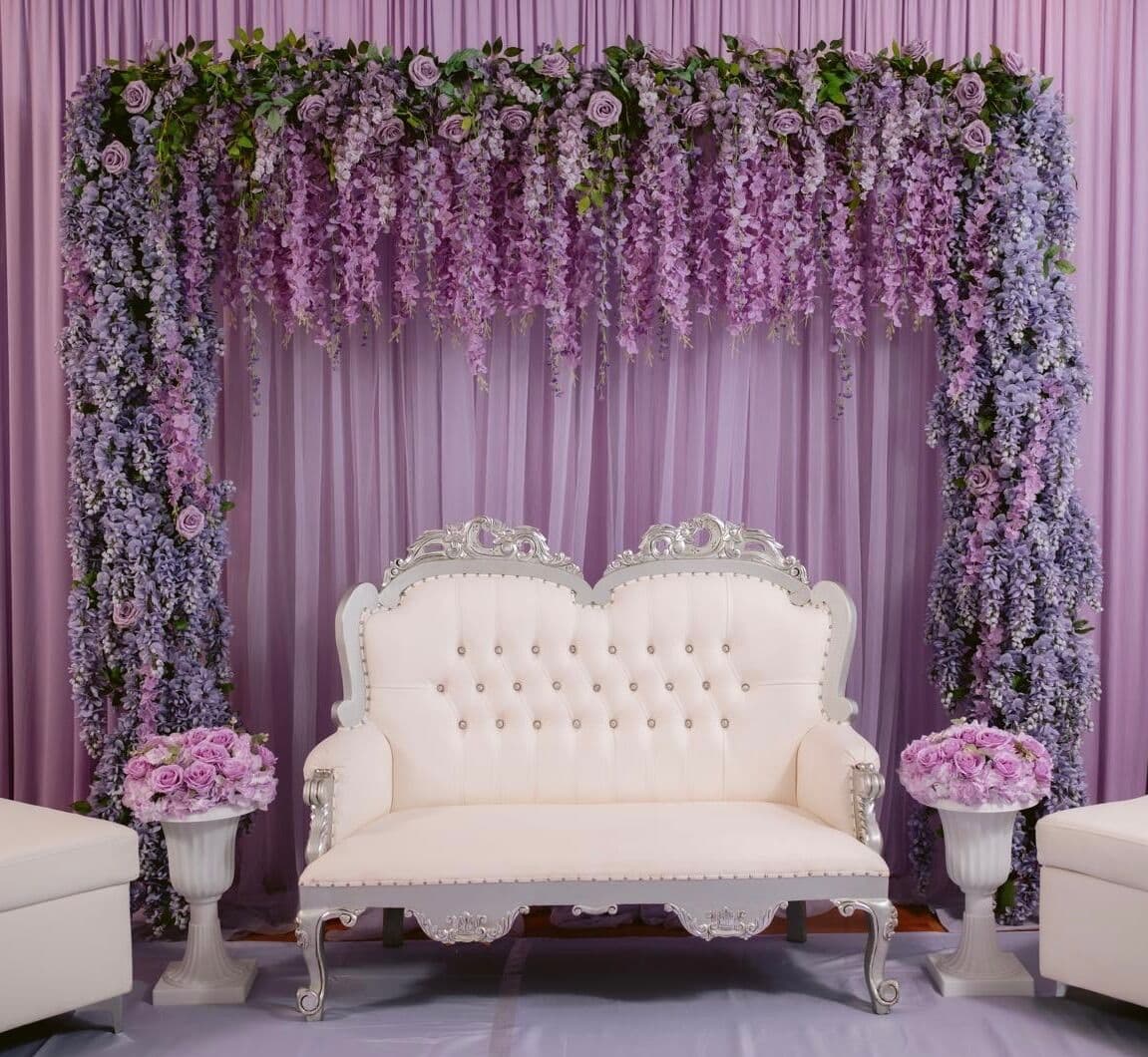 Engagement stage with a lush wisteria-style arch of cascading purple and lavender blooms over a lilac draped backdrop, framing an ornate white-and-silver tufted loveseat with lavender rose arrangements in white urns.