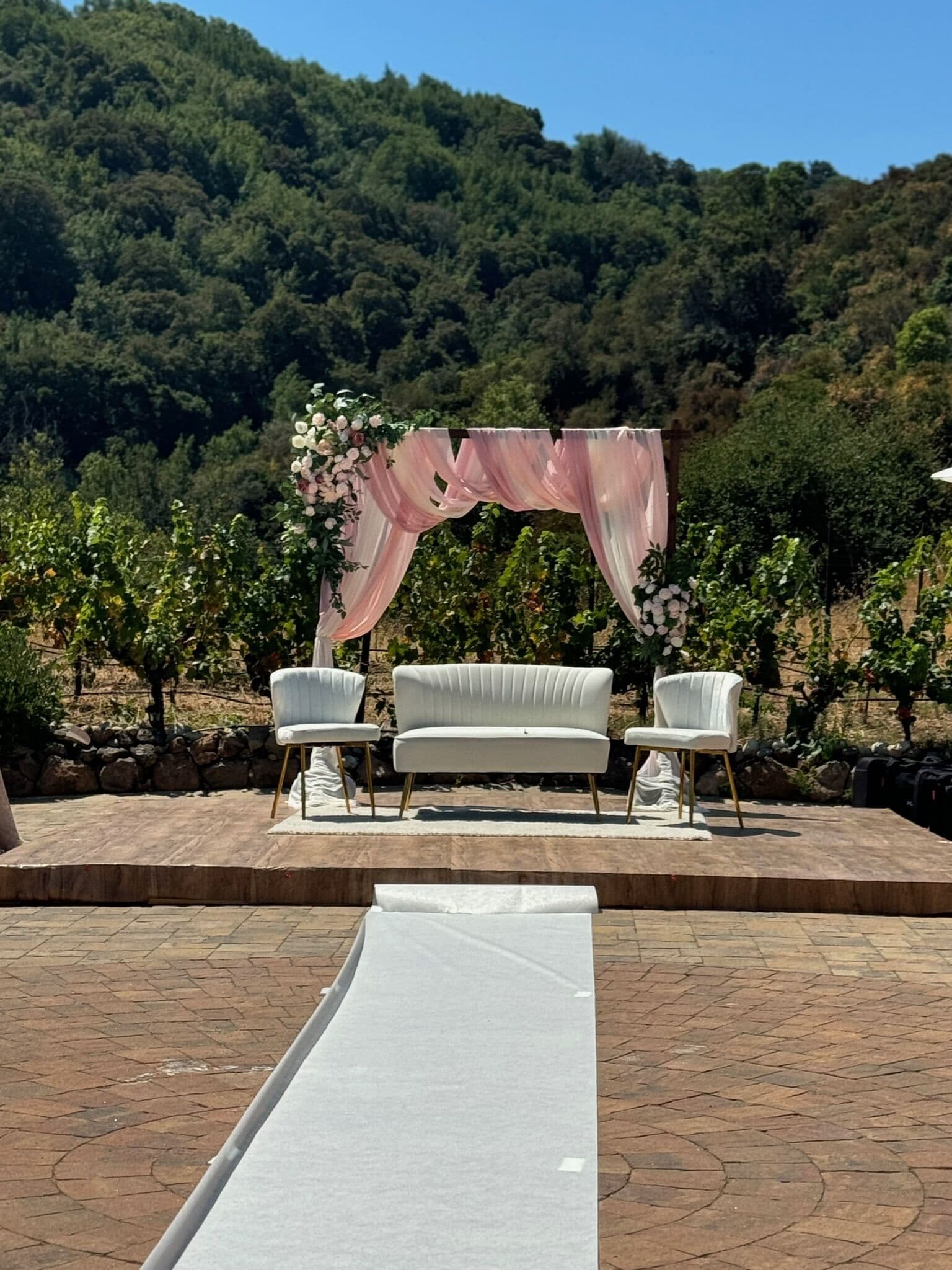 Outdoor Bay Area hillside ceremony stage with a blush chiffon-draped arch topped by white and pink roses, set with a channel-tufted white loveseat and two accent chairs on a wood platform overlooking a vineyard and green hills.