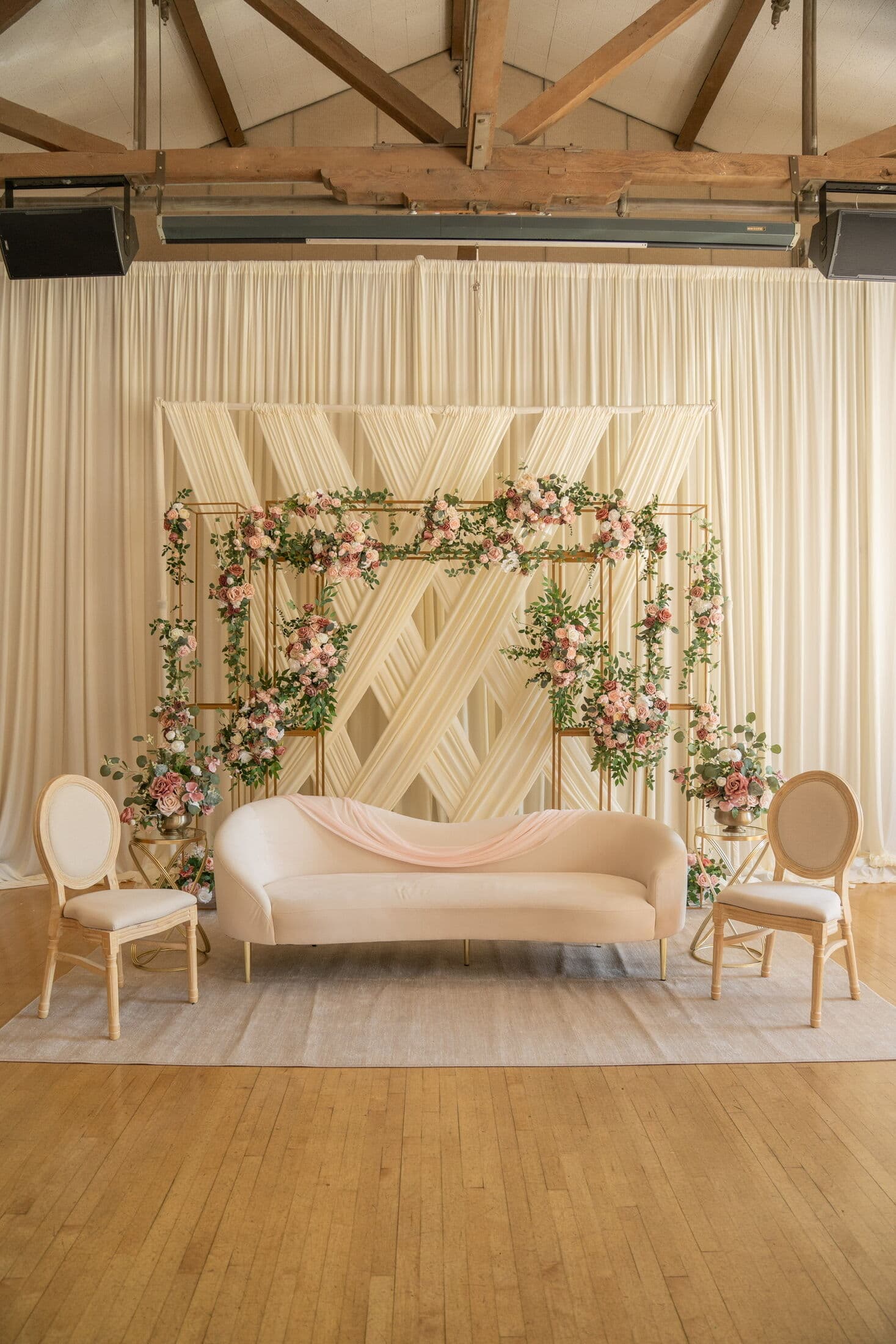 Barn-style wedding stage with exposed wood trusses, ivory pleated drapery and a geometric fabric backdrop framed by a gold trellis of blush roses and greenery, centered on a curved blush settee with a chiffon runner and oval-back chairs.