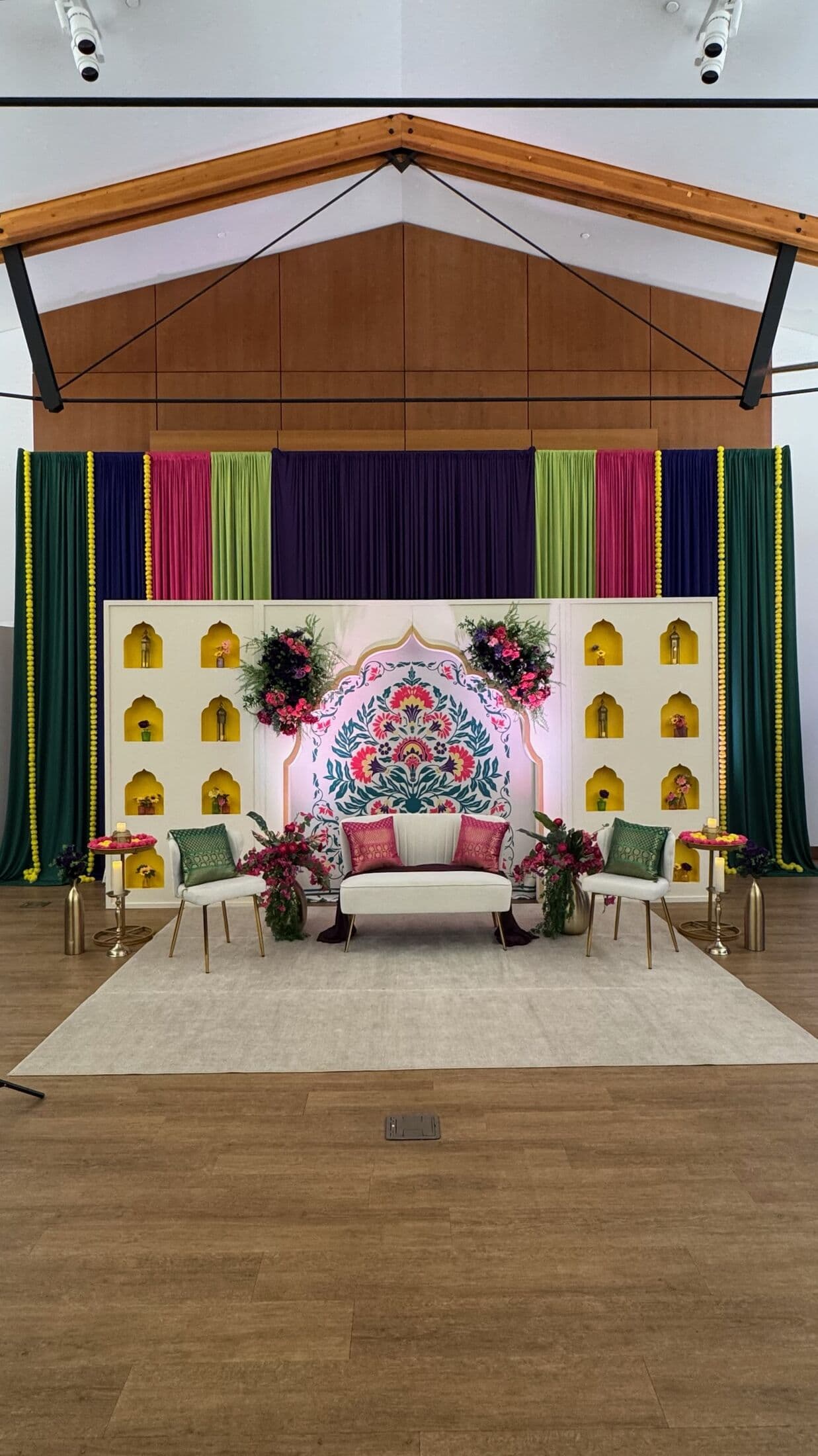Mehndi stage under wood beams with a hand-painted floral arch backdrop and ivory walls of gold-lit arched niches, framed by jewel-toned green, magenta and purple drapery with marigold garlands; a white settee with magenta and green cushions sits between accent chairs.