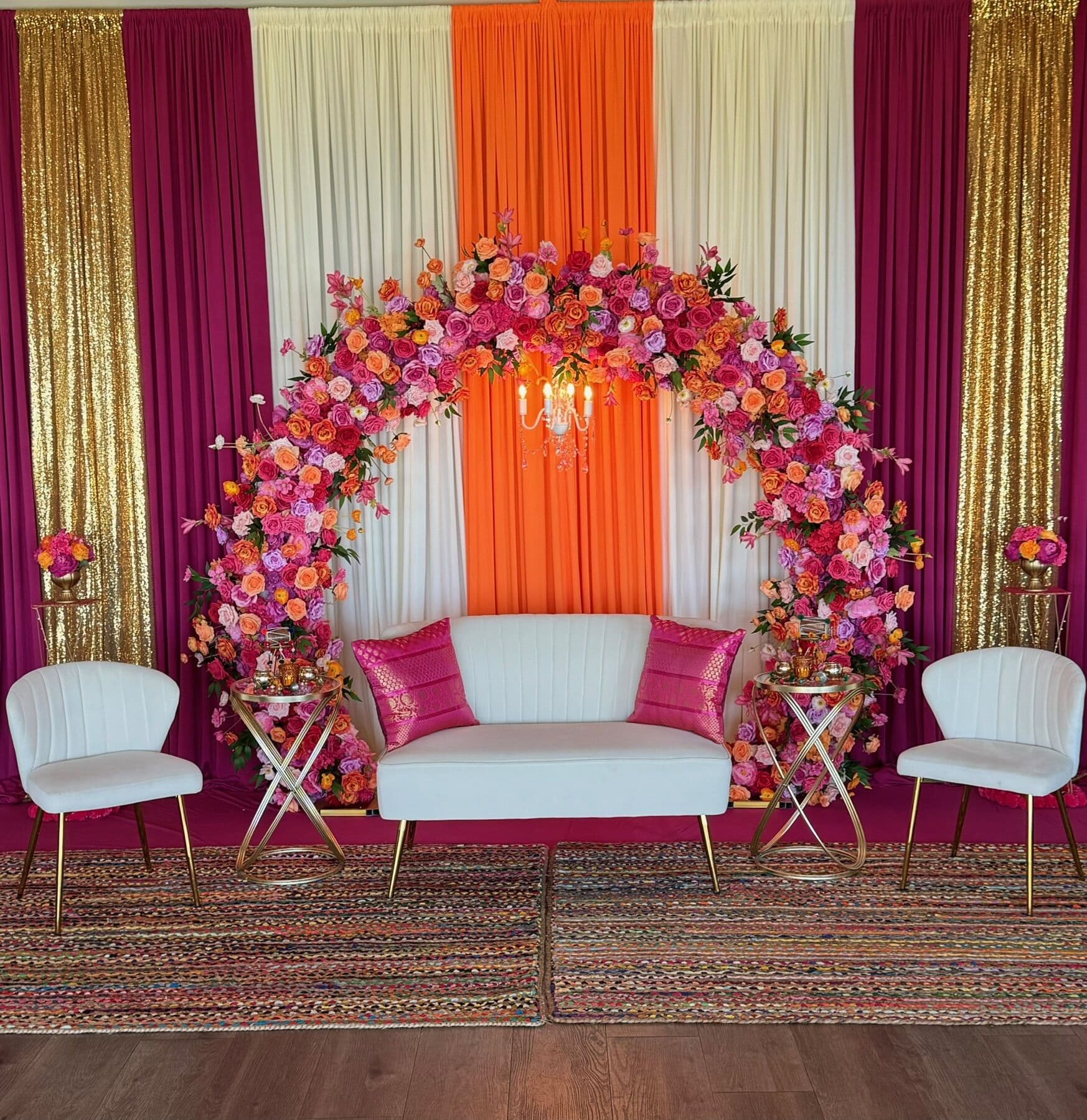 Mehndi stage with a full rose arch in pink, orange and magenta over drapery of magenta, ivory, orange and gold sequins, set with a white loveseat and magenta cushions between two accent chairs on layered woven rugs.