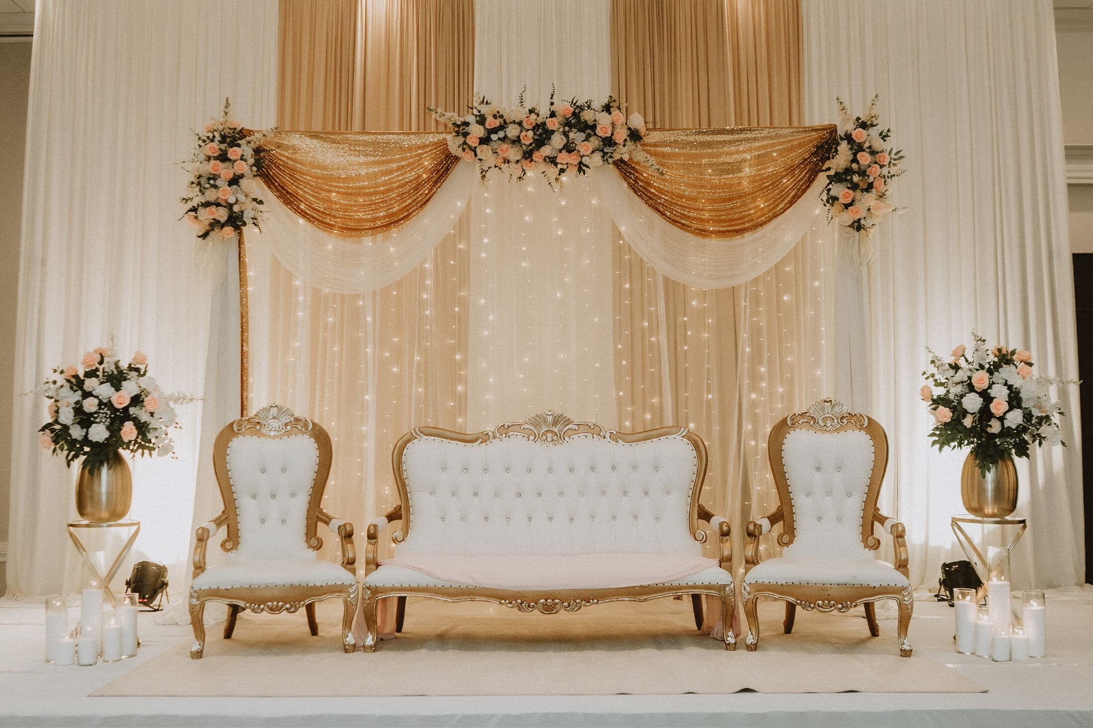 Walima stage by Memorable Moments by Ladle n Wok with champagne pleated and gold swag drapery over a twinkle-light curtain, blush floral toppers, and gold-framed white tufted thrones with a matching settee flanked by rose arrangements in gold urns.