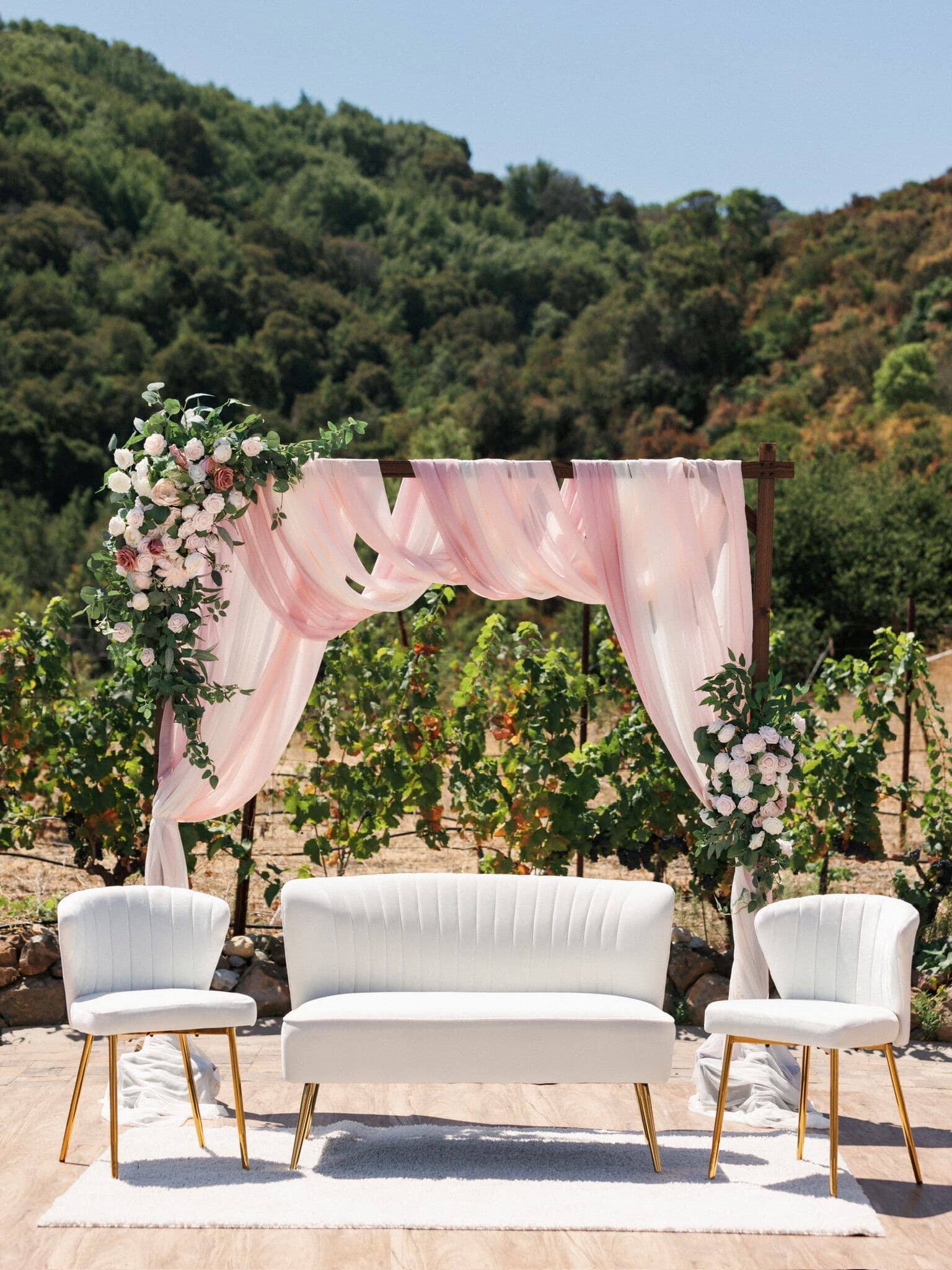 Outdoor Bay Area vineyard ceremony backdrop with a blush chiffon-draped arch accented by pink and white roses, set with a channel-tufted white loveseat and two gold-legged accent chairs on a wood platform against rolling green hills.