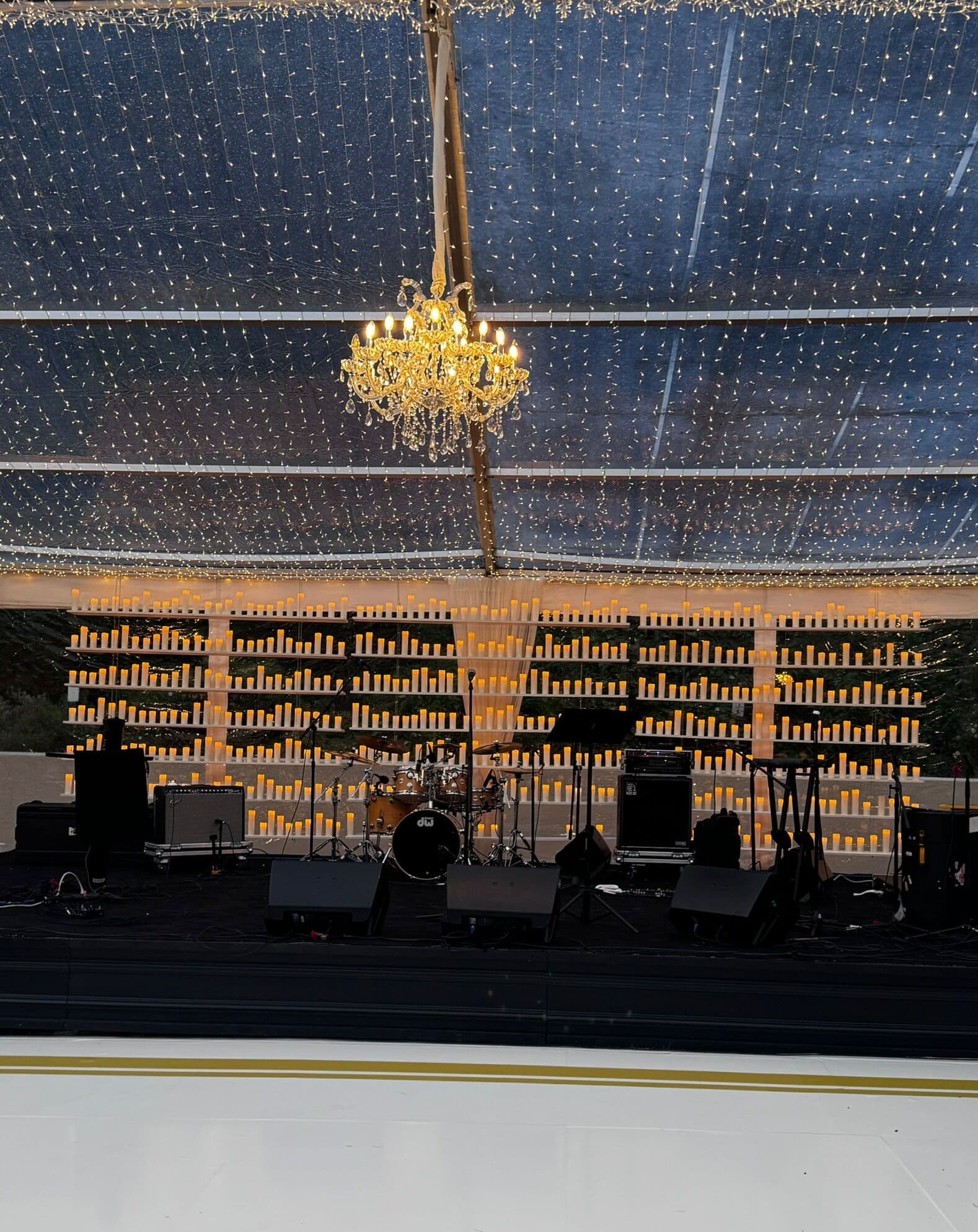 Clear-roofed tent stage lit by a warm crystal chandelier and twinkle-light ceiling, with a wall of glowing candles behind a live band's drum kit and equipment on a black stage.