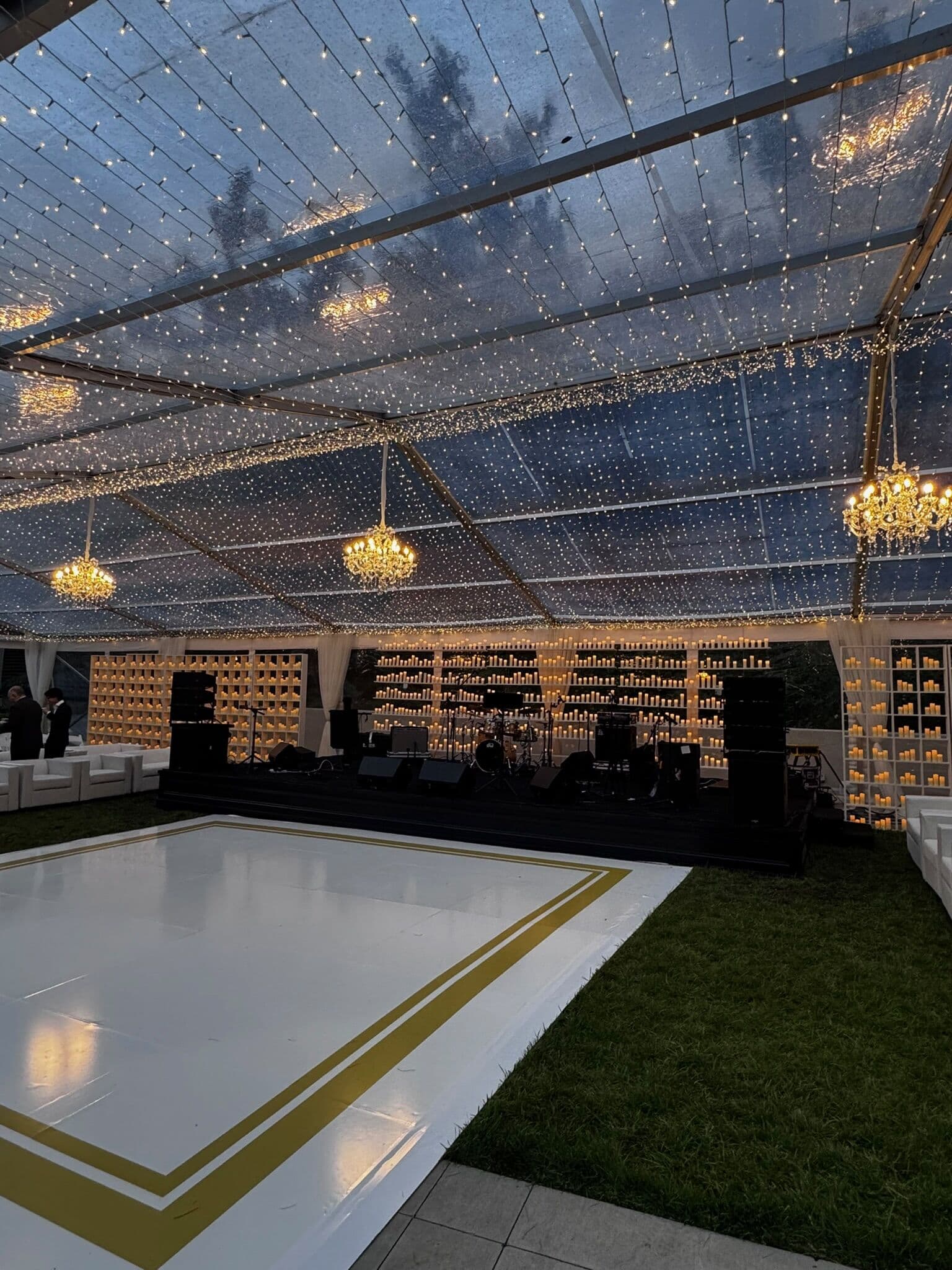 Twilight view of a clear-roof marquee with twinkle-light ceiling, hanging crystal chandeliers and a glowing candle wall behind the band, overlooking a white dance floor bordered in gold on the lawn.