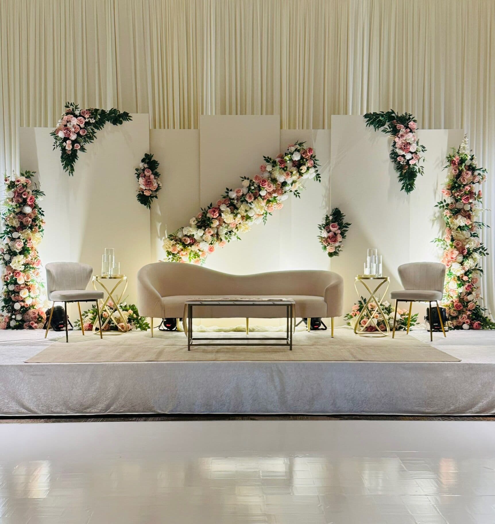 Wedding stage with paneled ivory backdrop lit warm, a diagonal garland of blush roses and greenery across the panels, corner floral columns and a curved beige sofa with a gold bench and accent chairs on a carpeted platform.