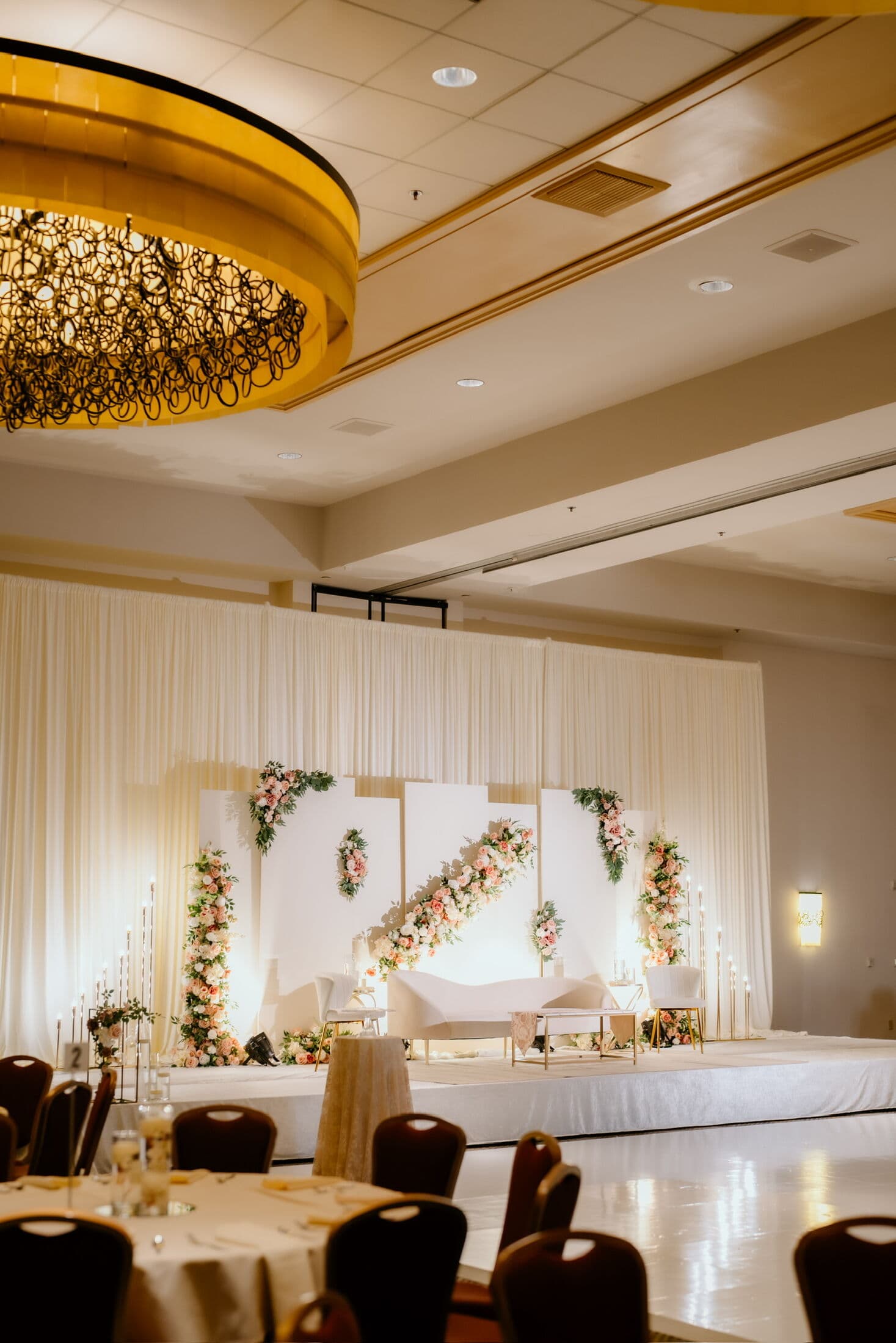 Ballroom walima stage with ivory drapery, white panels crossed by a diagonal blush rose garland and floral columns, lit warm behind a curved cream sofa and tall taper candles, viewed past round guest tables under a large gold ring chandelier.