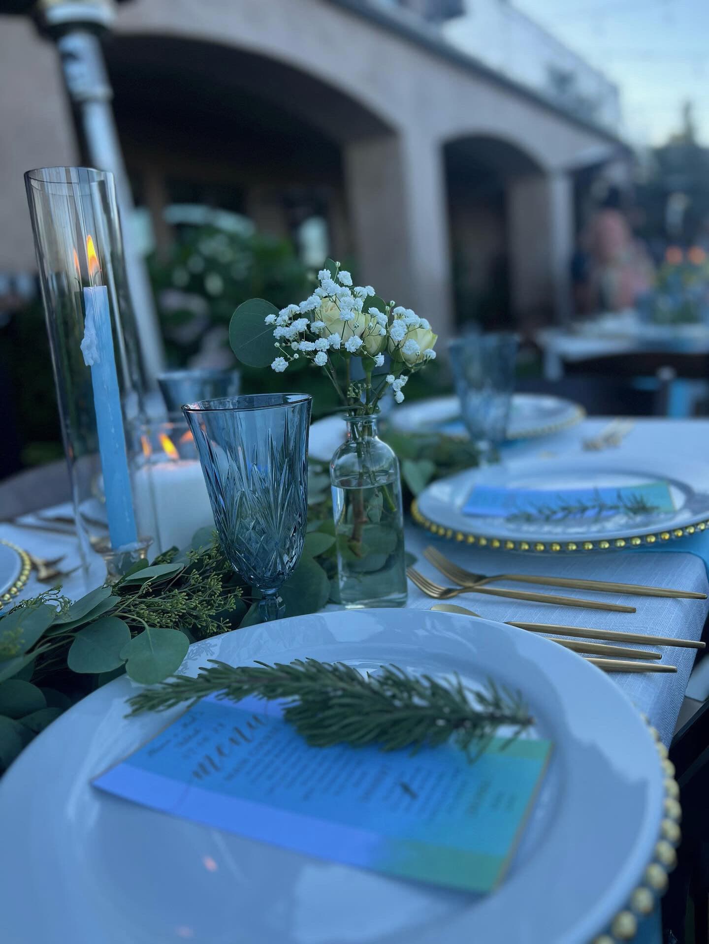 Outdoor Bay Area evening place setting with white plates on gold beaded chargers, gold flatware, cut-crystal blue goblets, taper candles in hurricane vases, a bud vase of white roses and baby's-breath, and eucalyptus running down the table.