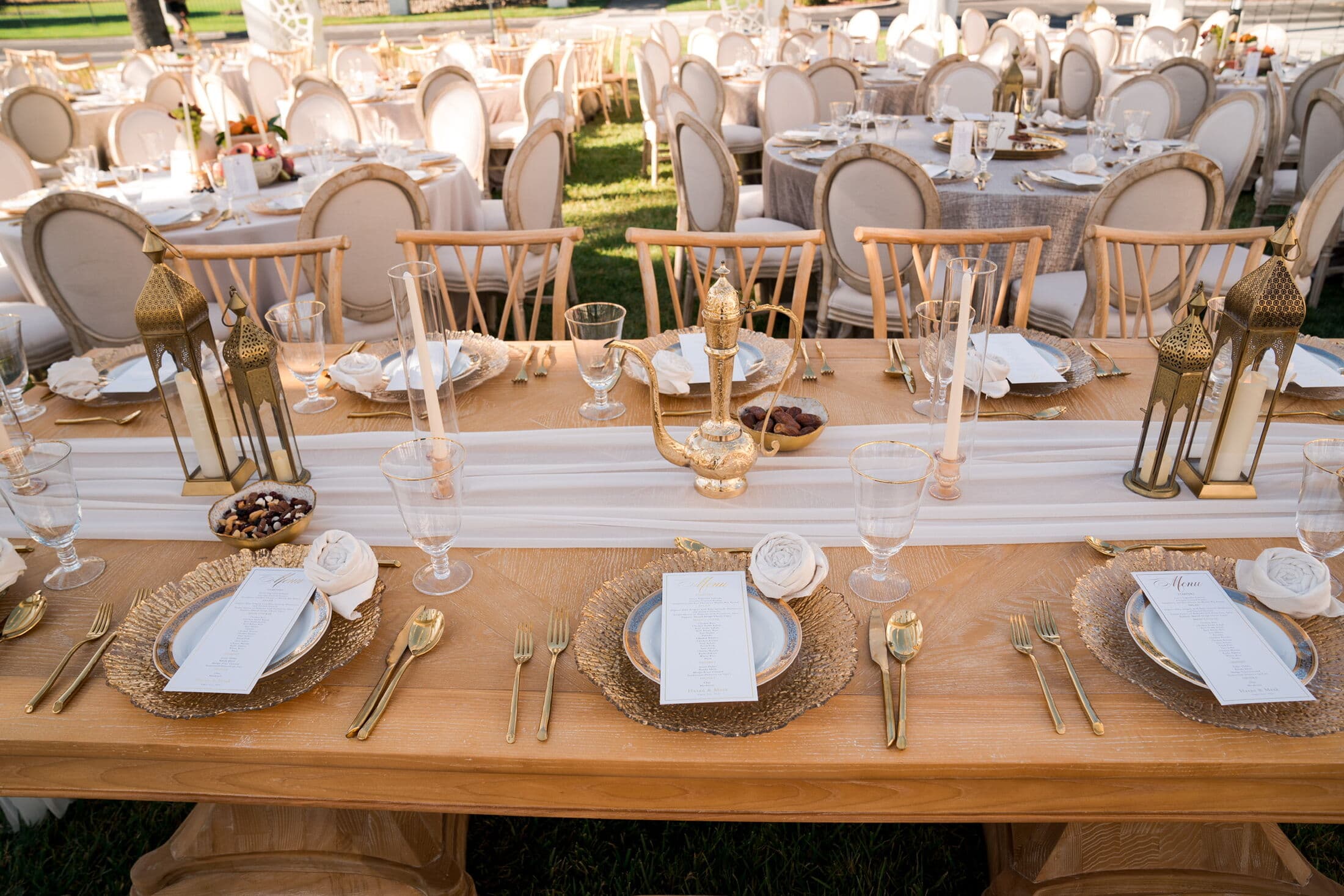 Long wooden banquet table with a white gauze runner, gold rattan chargers, gold flatware, printed menus, brass lanterns and a gold Arabic dallah coffee pot with a small bowl of dates at center, set outdoors on a lawn by Memorable Moments by Ladle n Wok.