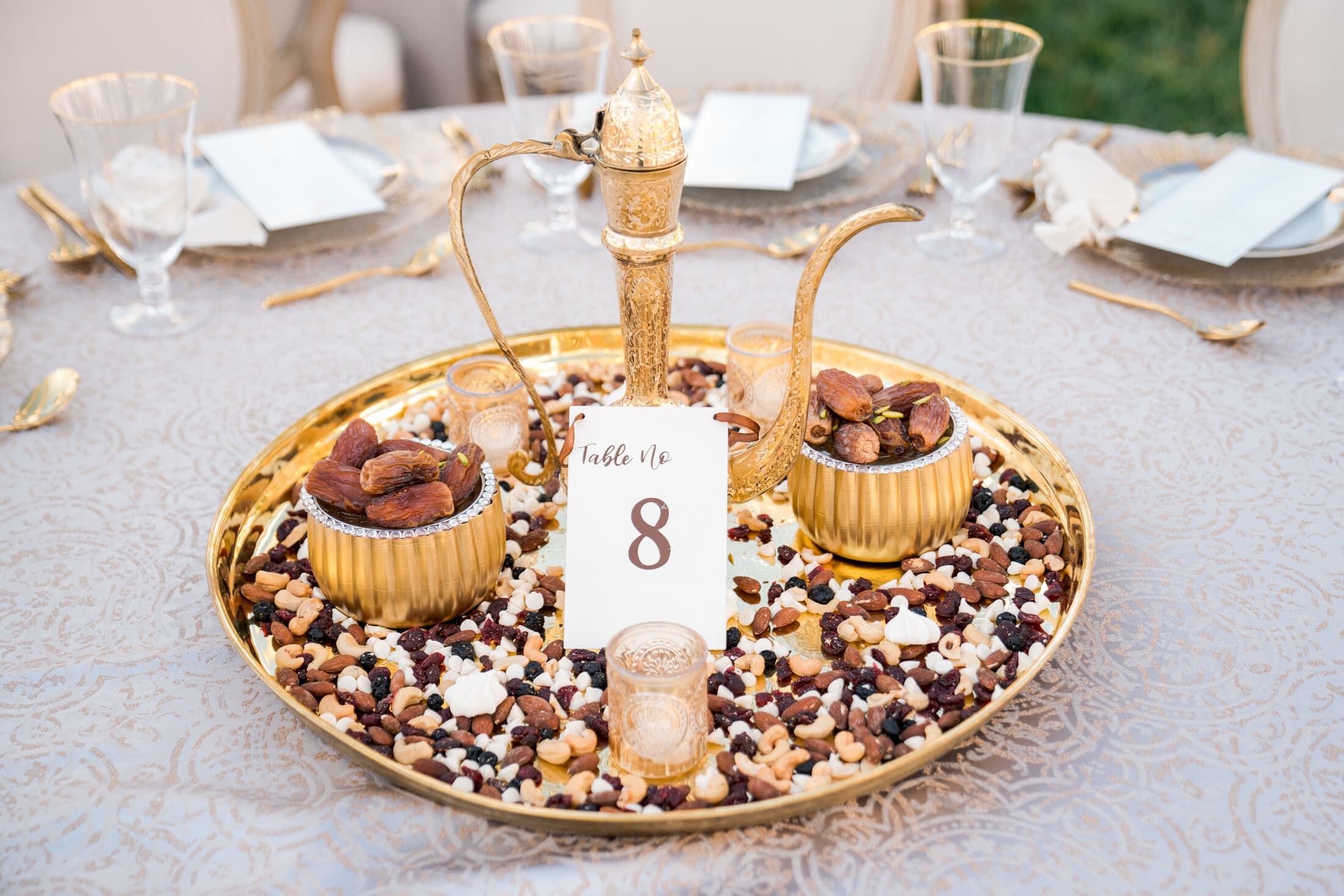 Round gold tray centerpiece filled with mixed nuts, dried fruit and dates in footed brass bowls flanking a gold dallah coffee pot, with a white 'Table No 8' card and votive candle, styled for a religious celebration.