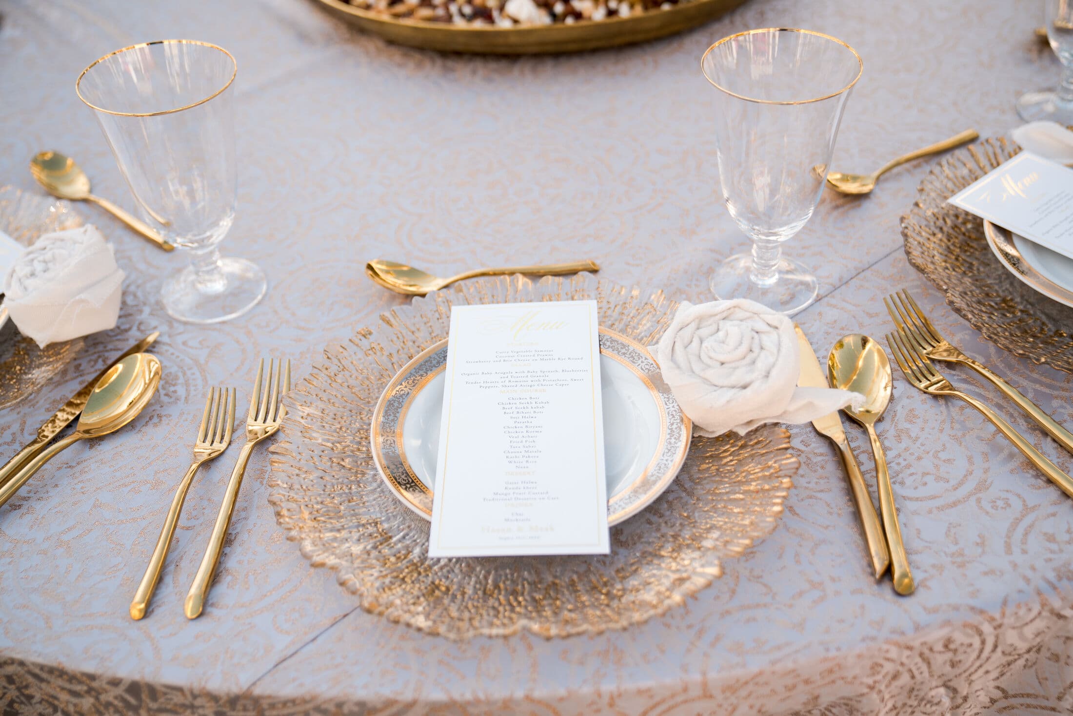 Place setting on a pale blue damask linen with a textured gold glass charger, gold-rimmed china, a printed menu, a fabric rosette napkin, gold-rimmed goblets and gold flatware, part of a Bay Area wedding tablescape.