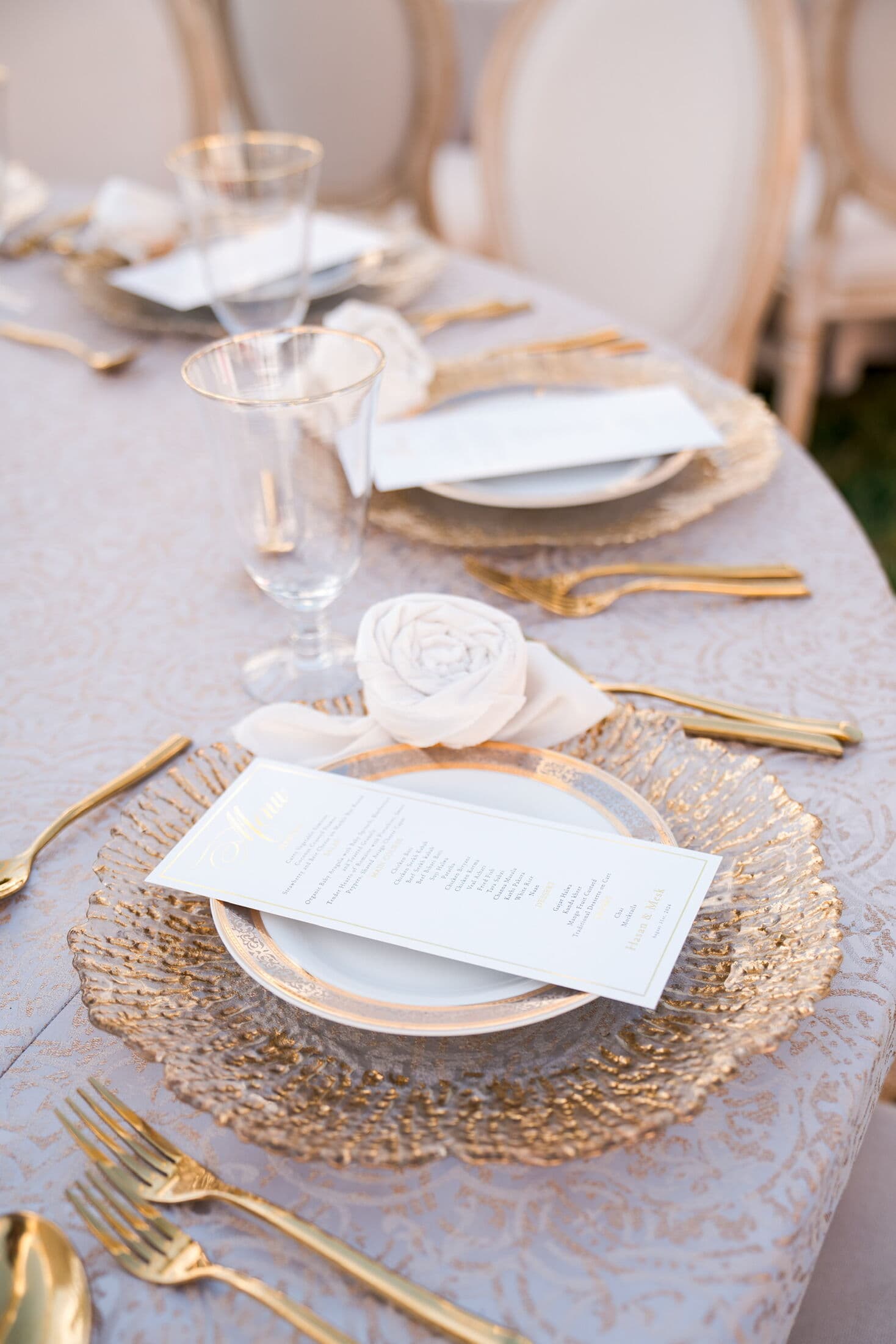 Oval table in silver-gold brocade linen set with gold-flecked glass chargers, gold-rimmed plates, printed menus, twisted chiffon rosette napkins, gold-rimmed goblets and gold flatware beside cushioned oval-back chairs.