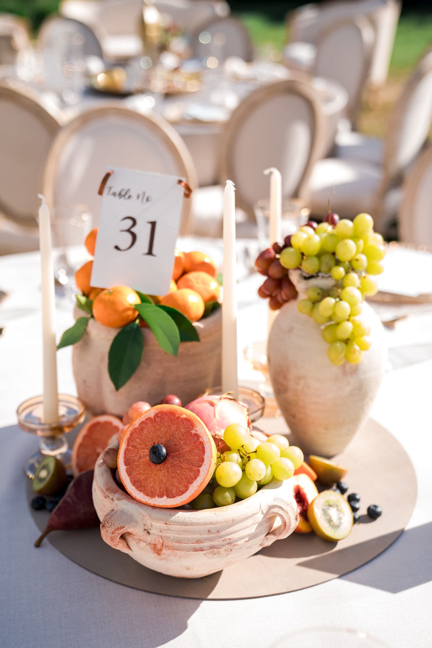 Mediterranean-style fruit centerpiece in weathered terracotta urns holding clementines, green grapes and figs with a halved blood orange, kiwi and blueberries, ivory taper candles and a 'Table No 31' card at an outdoor Bay Area reception.