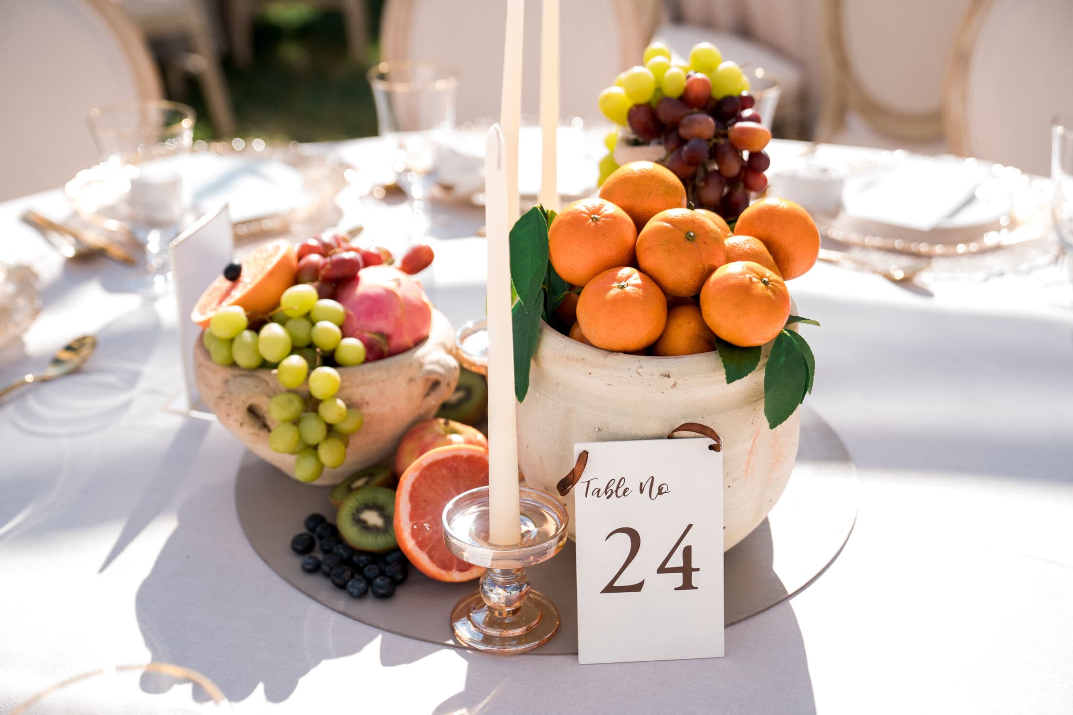 Rustic ceramic urns overflowing with oranges, green and red grapes, dragon fruit and halved citrus with kiwi and blueberries, an ivory taper candle and a 'Table No 24' card on a round white-linen table outdoors.