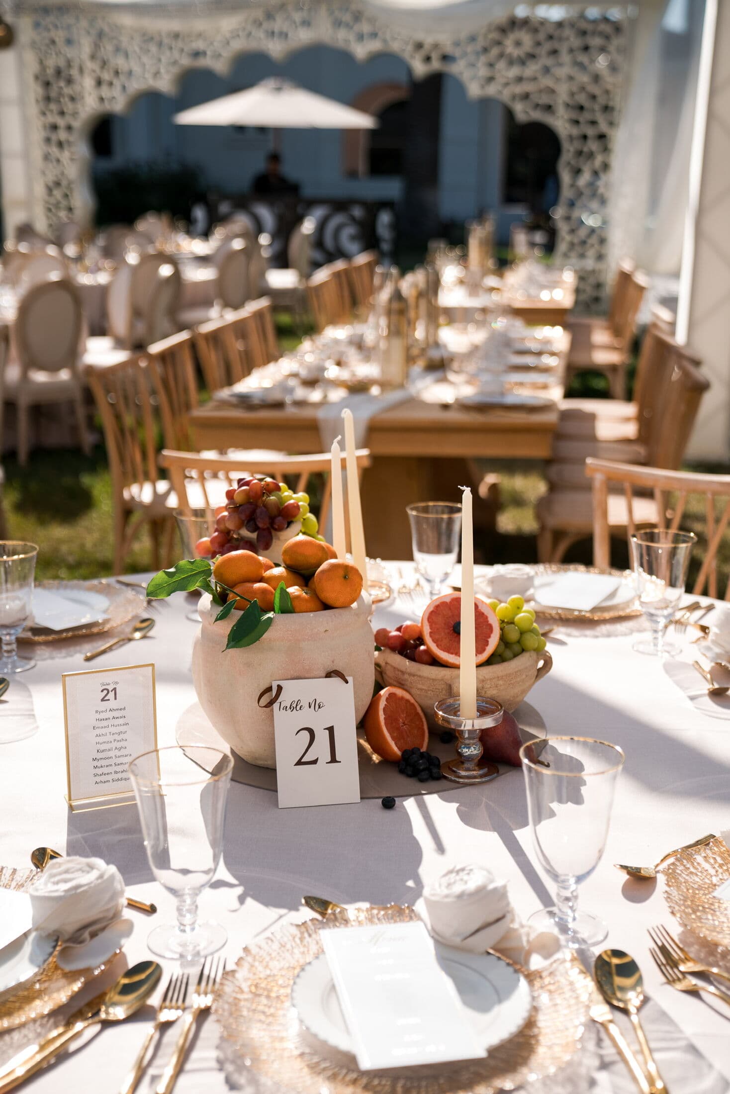 Round guest table under a tent with white linens, gold woven chargers, folded napkins, gold flatware and a terracotta-urn fruit centerpiece of oranges, grapes and citrus with a 'Table No 21' card by Memorable Moments by Ladle n Wok.