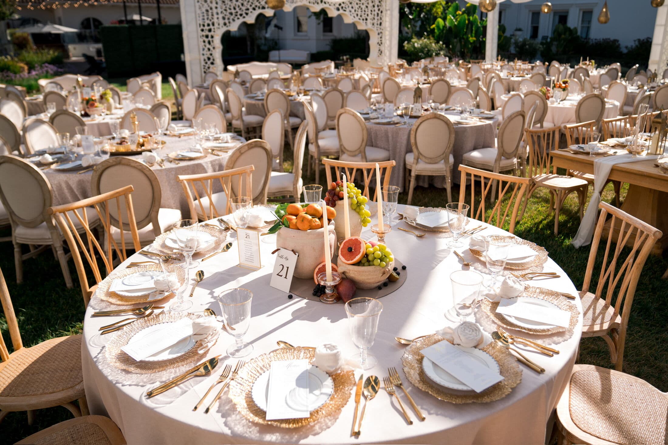Round white-linen reception table with gold woven rattan chargers, gold flatware and a low fruit-and-candle centerpiece, set among rows of tables on a sunlit Bay Area lawn.