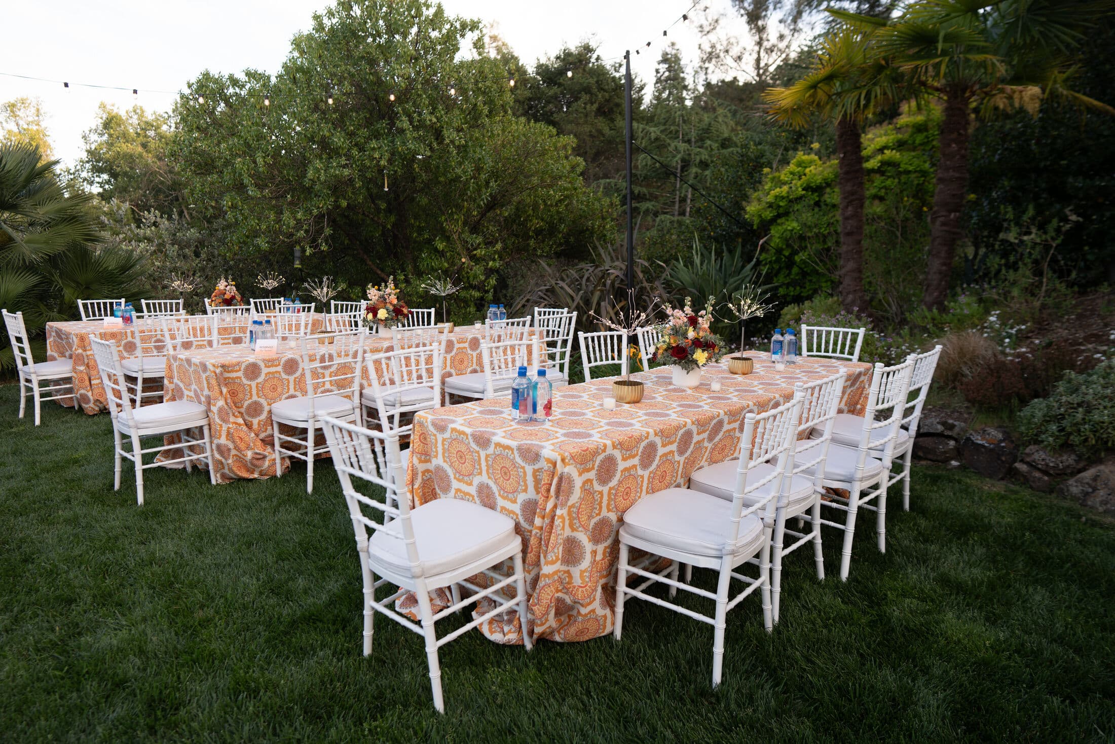Outdoor garden tables dressed in orange and cream medallion-print linens with white Chiavari chairs, low floral centerpieces and blue water bottles, set on grass beside greenery and string lights.