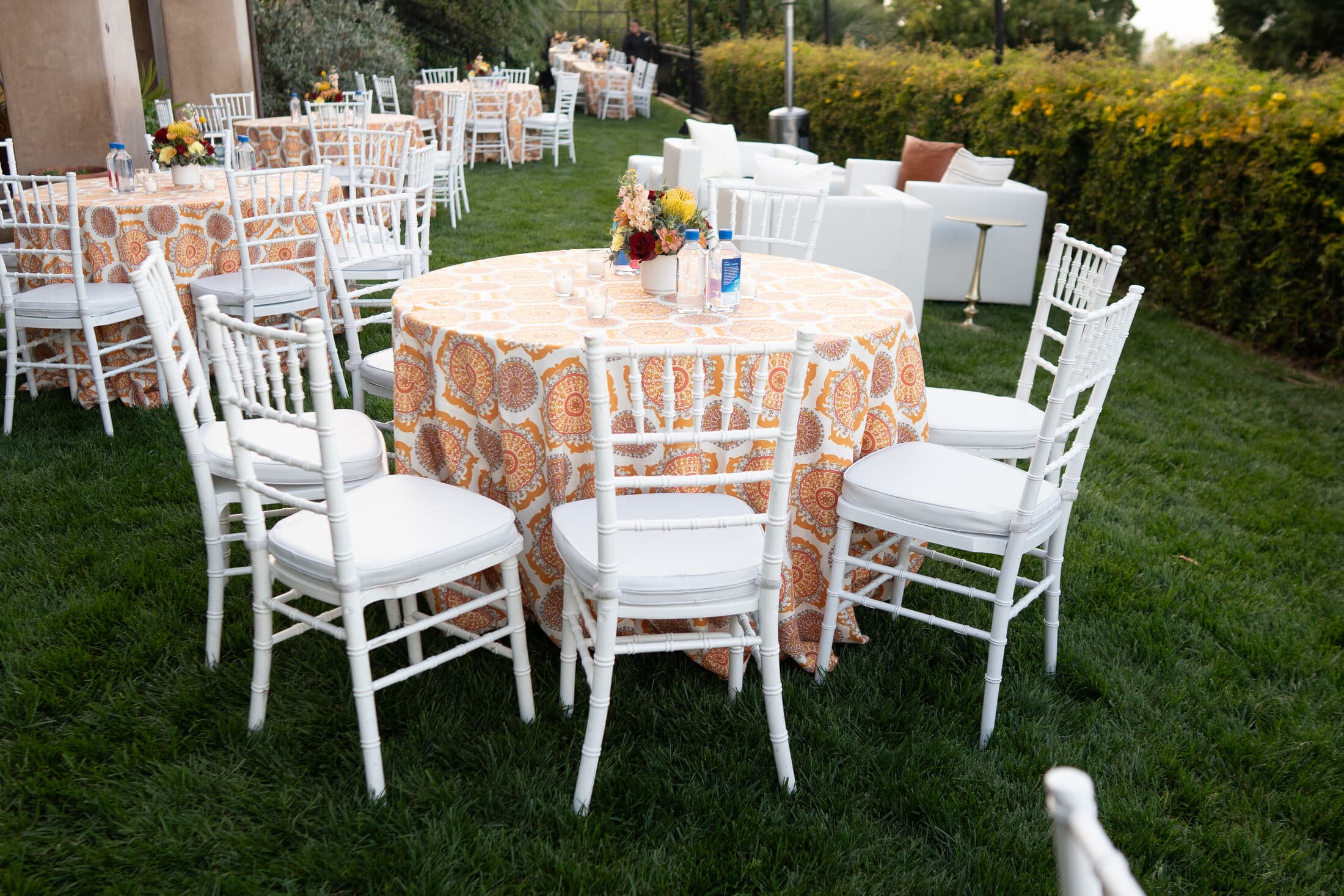 Round table in an orange and cream medallion-print linen ringed by white Chiavari chairs, topped with a bright floral centerpiece and glassware, on a lawn beside a white lounge sofa.