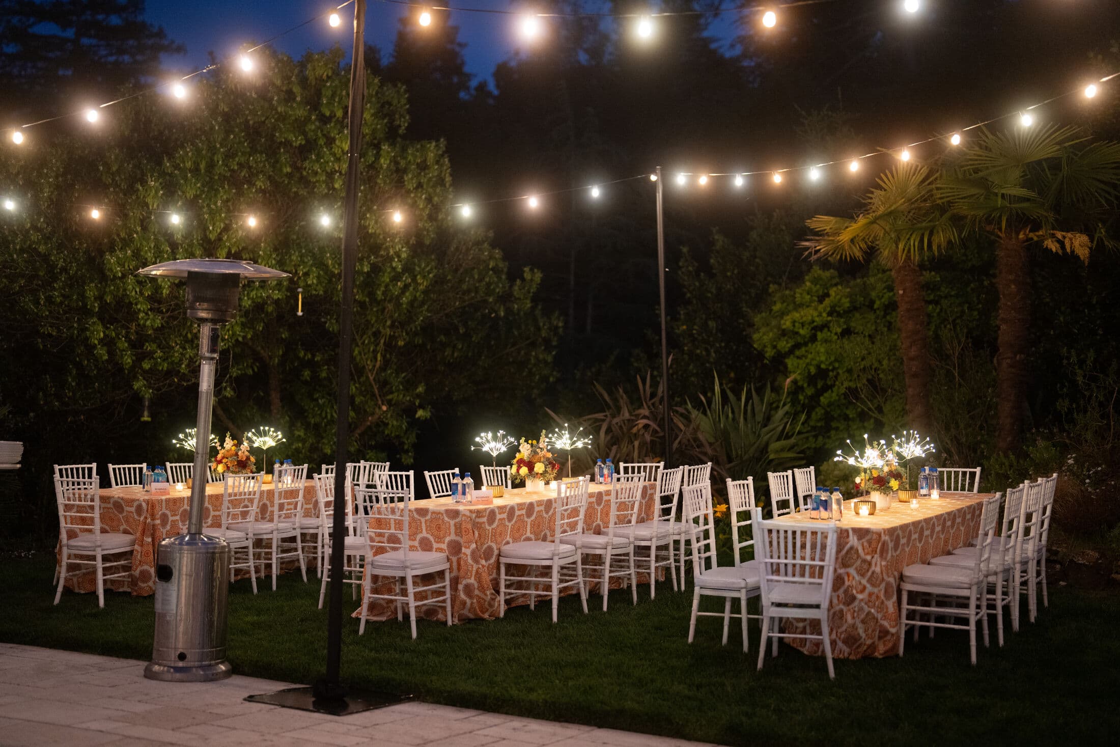 Nighttime garden reception with U-shaped banquet tables in orange medallion-print linens, white Chiavari chairs, candlelit centerpieces and a patio heater, glowing under canopies of string lights.