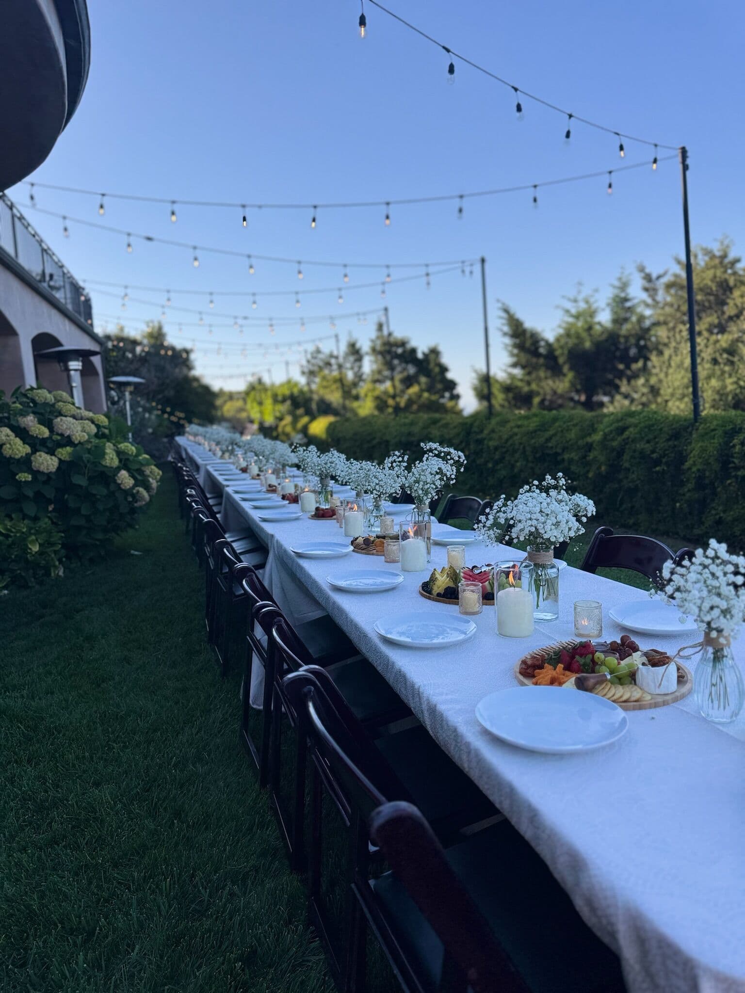 Long white-linen banquet table set outdoors under string lights with white plates, votive candles, bud vases of baby's breath and wooden grazing boards of fruit, cheese and charcuterie, lined with dark folding chairs at dusk in the Bay Area.