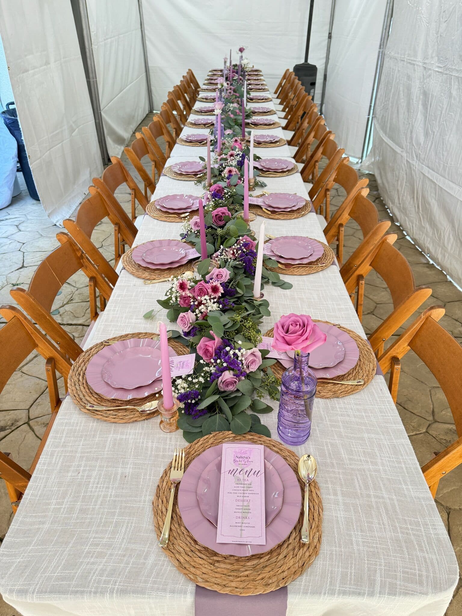 Long tented table in ivory textured linen with woven rattan chargers, mauve plates, pink and lavender taper candles, gold flatware, purple menu cards and a lush garland of pink roses, purple blooms and eucalyptus down the center.